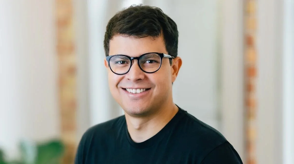 A young man with dark hair, wearing glasses and a black t-shirt, smiling at the camera indoors.