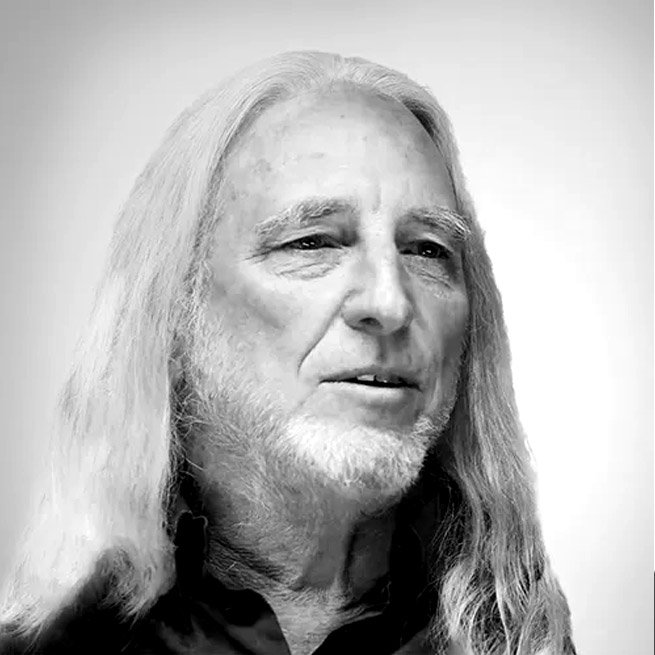 Black and white portrait of an older man with long, gray hair and a beard, wearing a dark shirt.