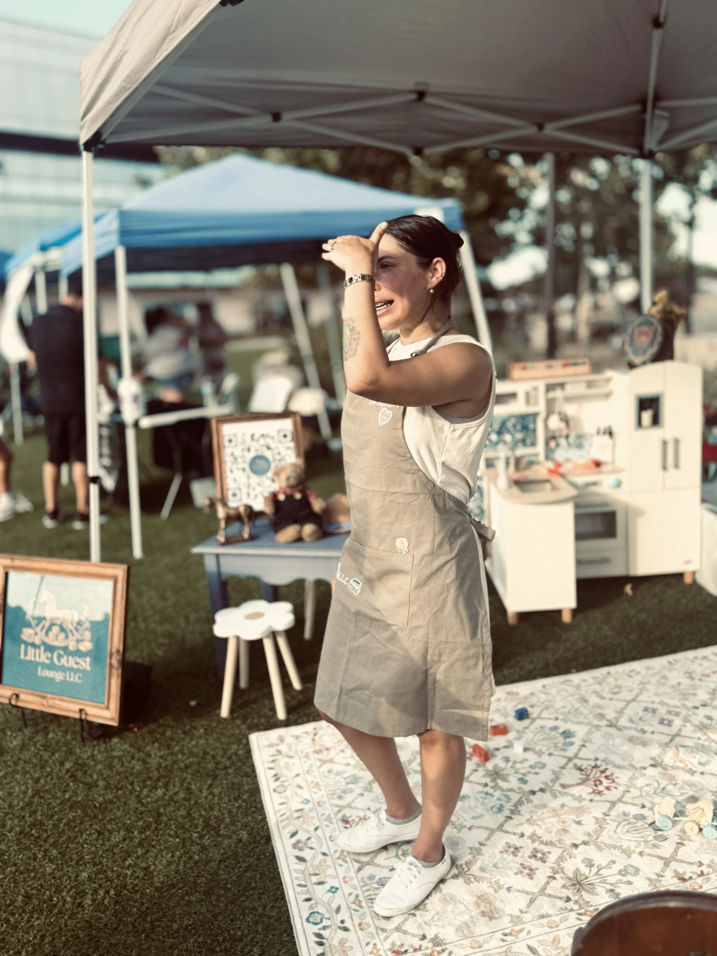 A woman with dark hair, freckles, and tattoos on her left arm is standing outdoors under a canopy. She is touching her forehead with her left hand and appears to be talking or reacting. She is wearing a beige apron over a sleeveless shirt and white s