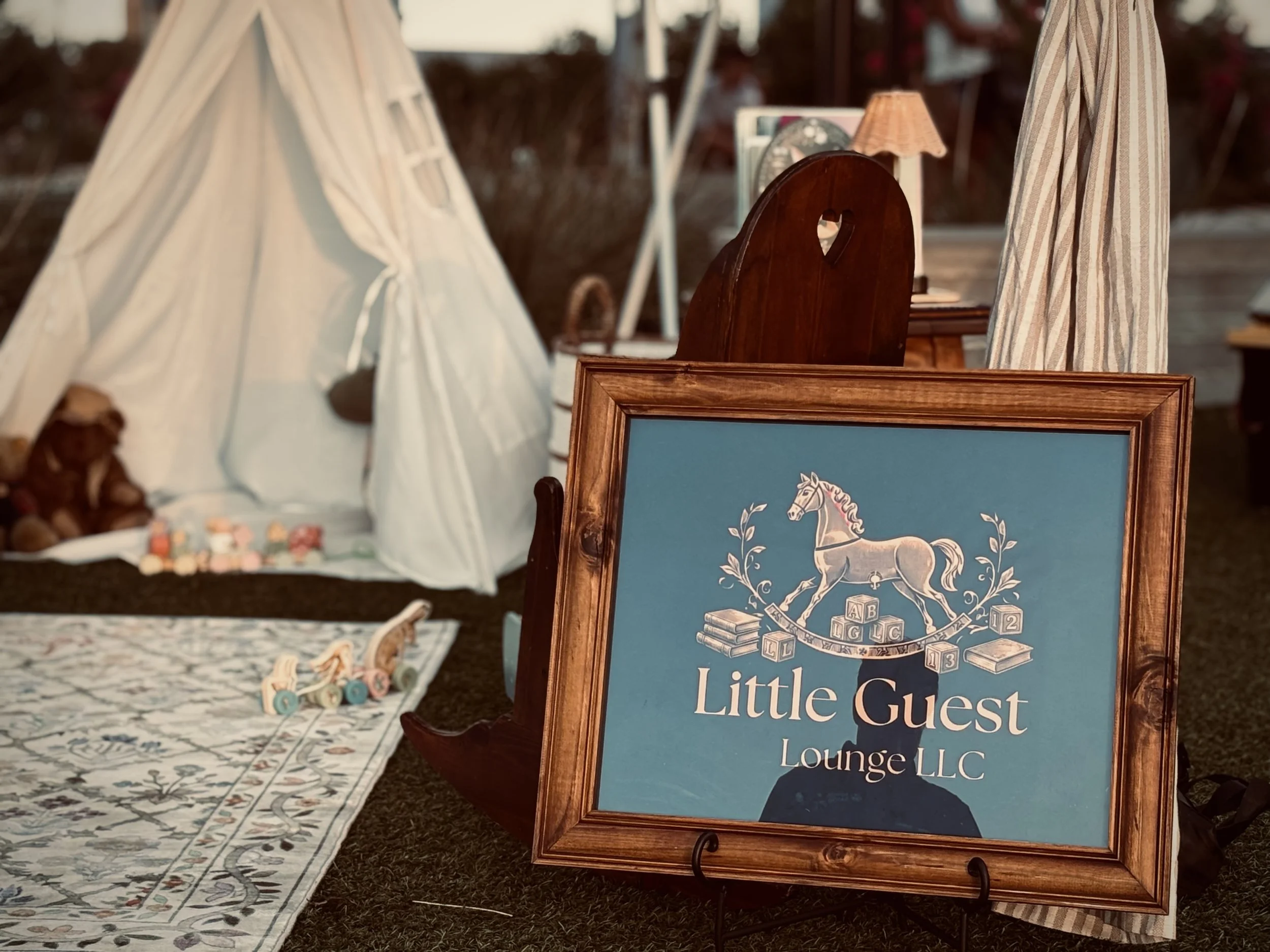 A framed sign for Little Guest Lounge LLC sits on an easel at an outdoor event. In the background, there is a white teepee tent and a table with decorative items and toys.