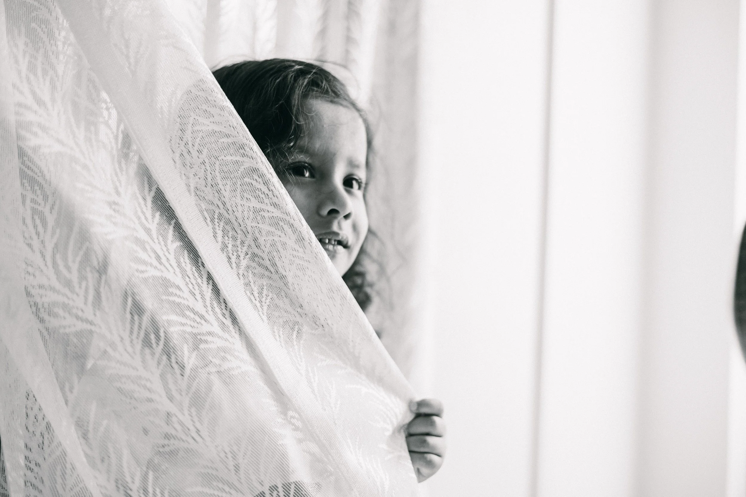 A young girl with dark hair peeking from behind a lace curtain, partially looking out a window on a bright day, holding a white curtain with her right hand.