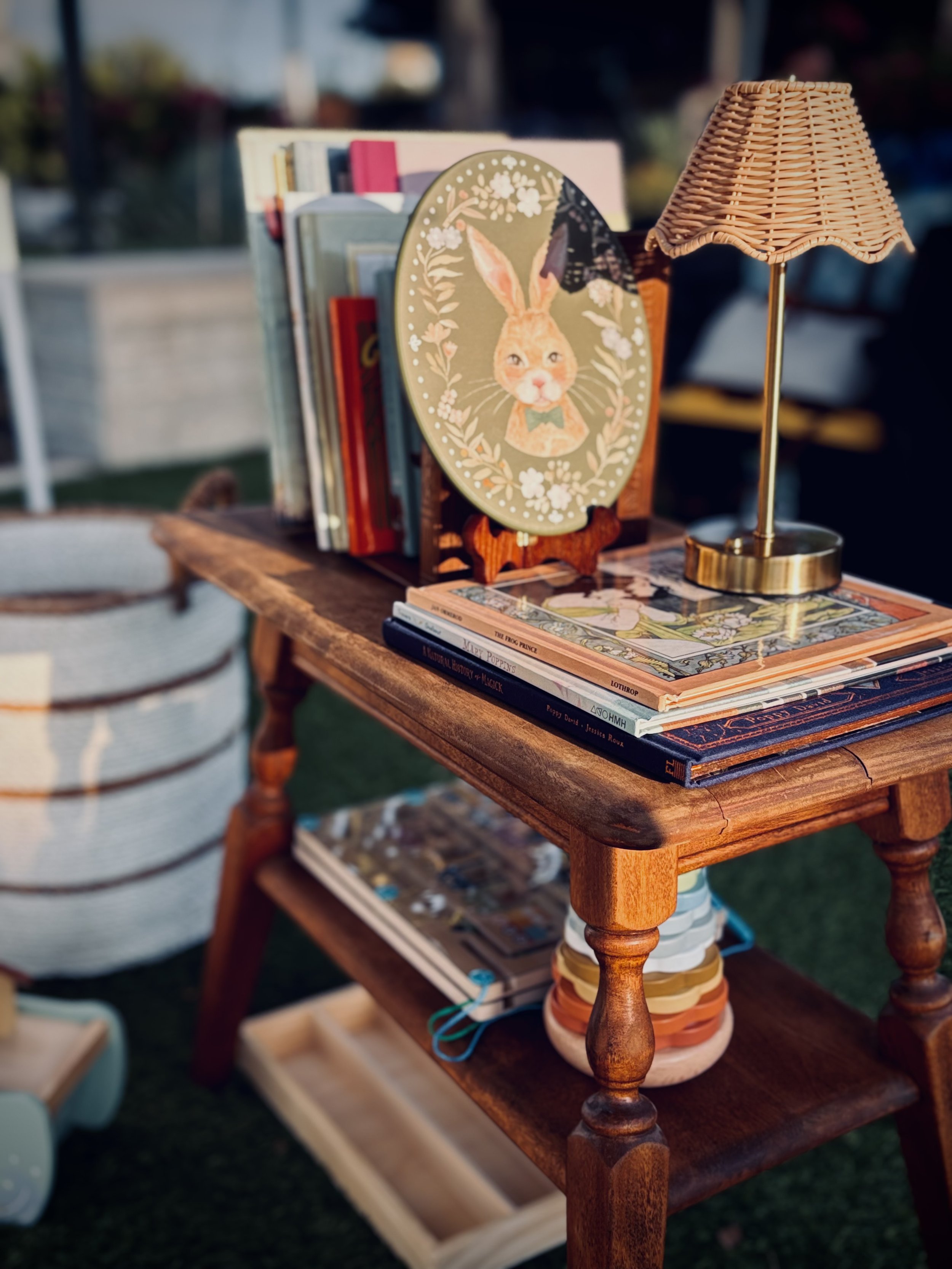 A wooden table with books, a decorative bunny picture, and a wicker lampshade at an outdoor market.