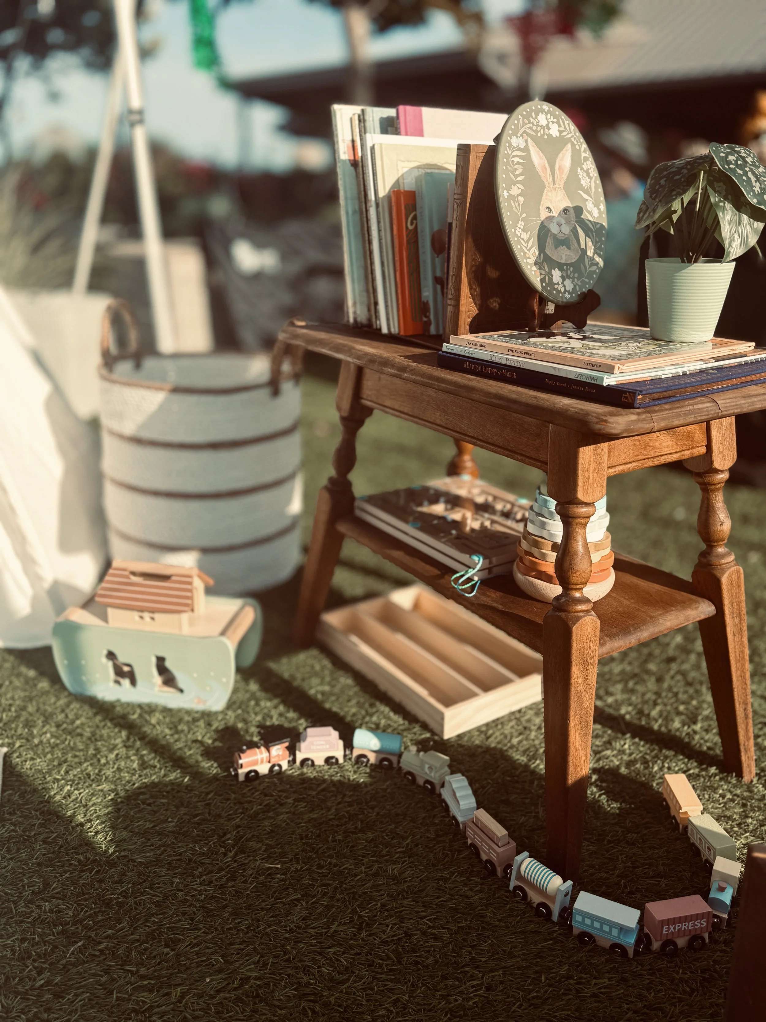 A small wooden table with books, a decorative plate with a rabbit illustration, a potted plant, and several children's toy trains on a grassy surface.