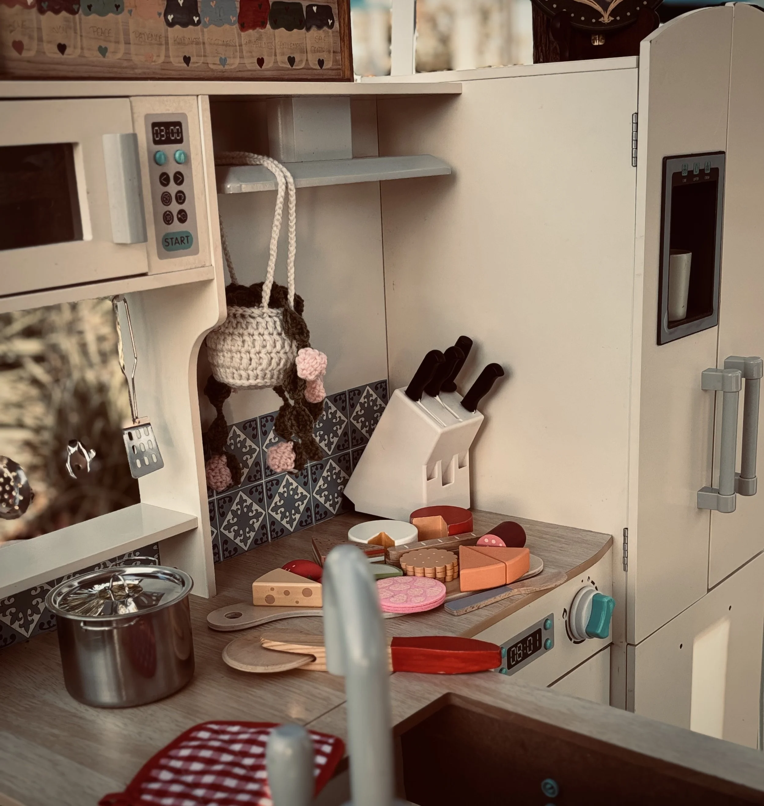 A children's play kitchen with various toy food items and utensils on the wooden countertop and shelves. The play kitchen features a digital clock, stove, and oven, with a small hanging basket and decorative elements. The scene is part of a cozy, wel