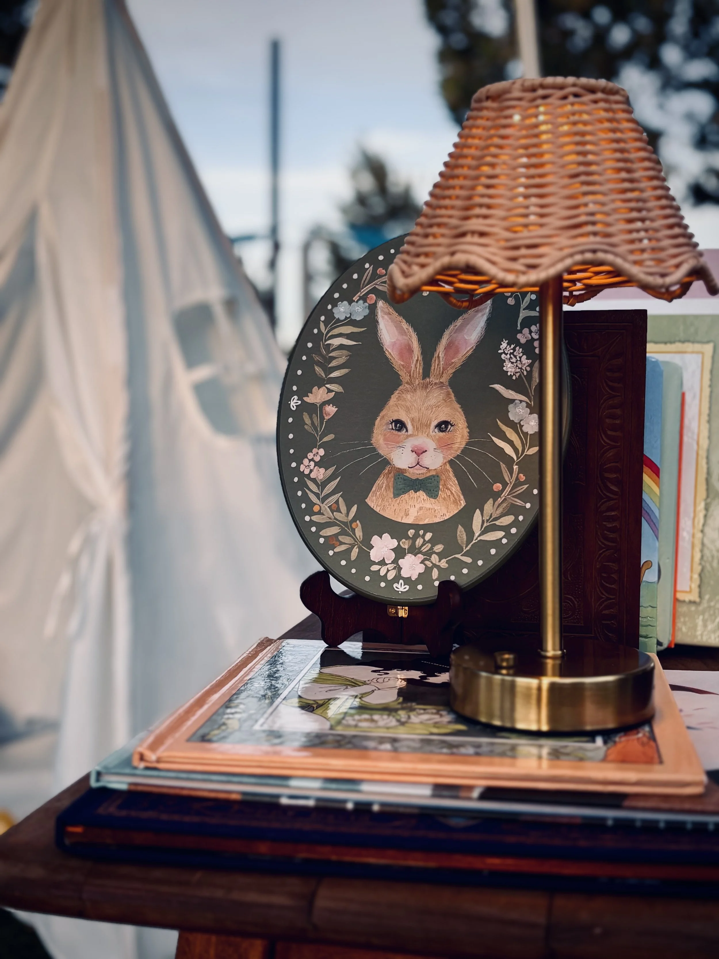 Decorative rabbit-themed framed plaque on a table, with a table lamp and books nearby, a tent visible in the background.