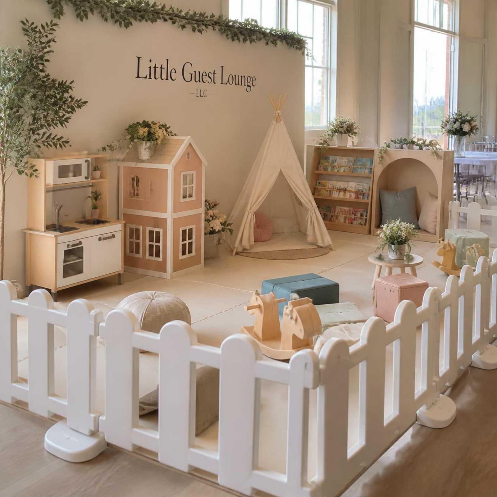 Decorated children's play area with a white picket fence, mini kitchen, playhouse, teepee, bookshelf, and seating with flowers, in a bright room with large windows.