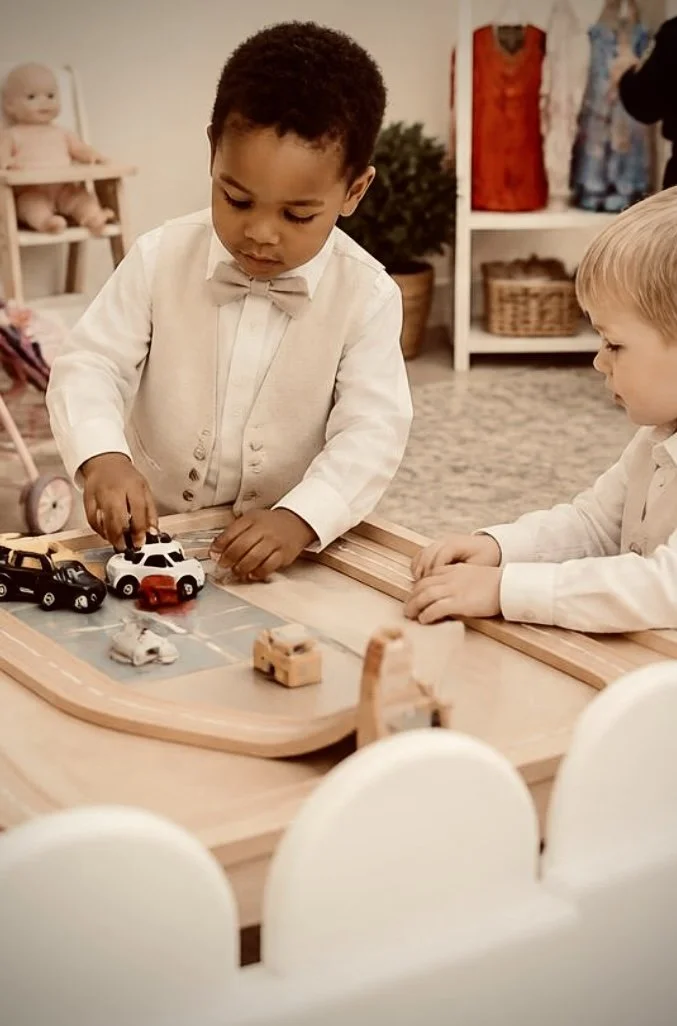 Two young boys dressed in formal clothes playing with toy cars on a wooden race track in a room with shelves containing toys and clothes.