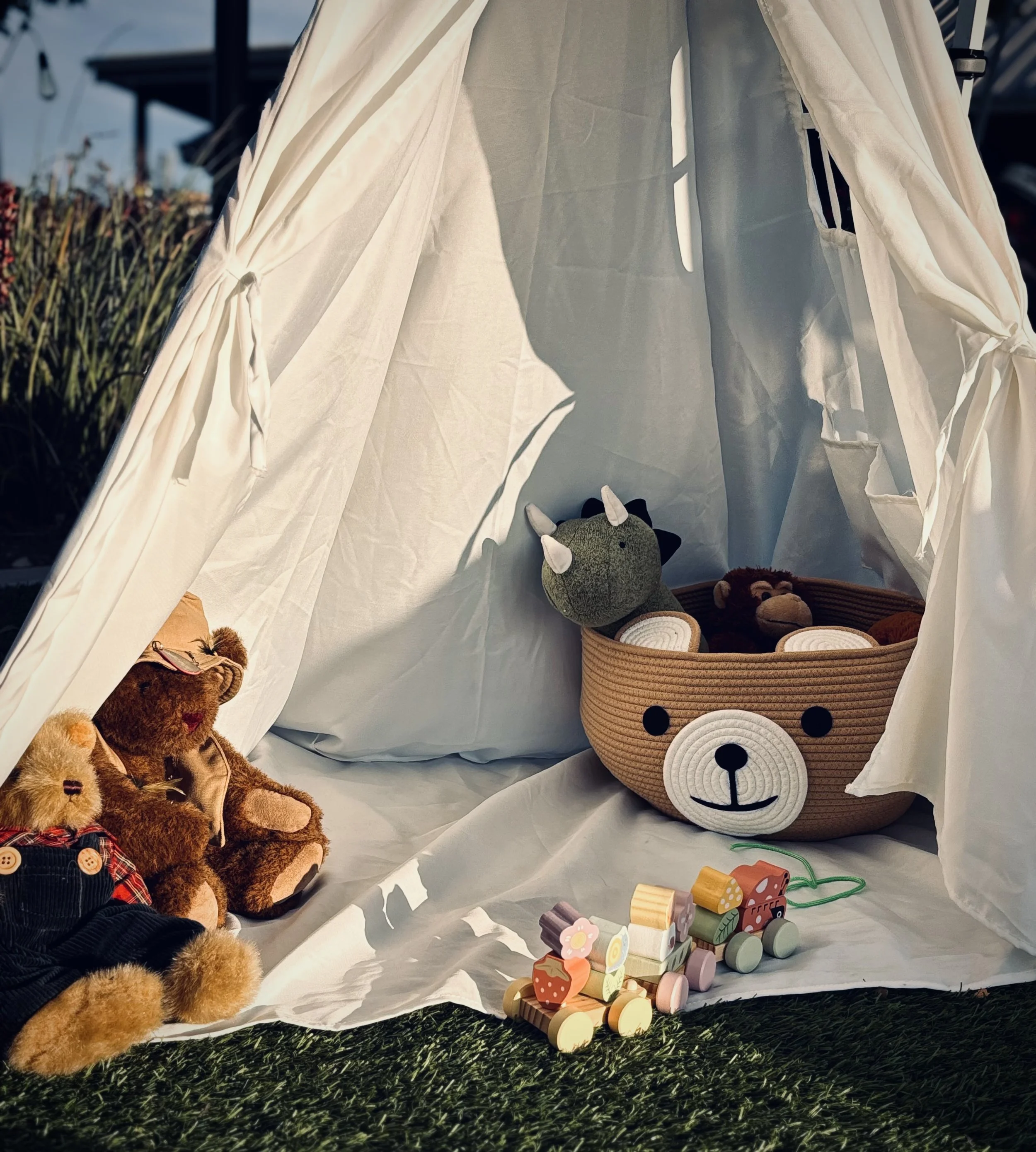 Children's toys inside a white fabric play tent, including plush animals, wooden toy blocks, and a basket with plush teddy bears, set on a green artificial grass surface.