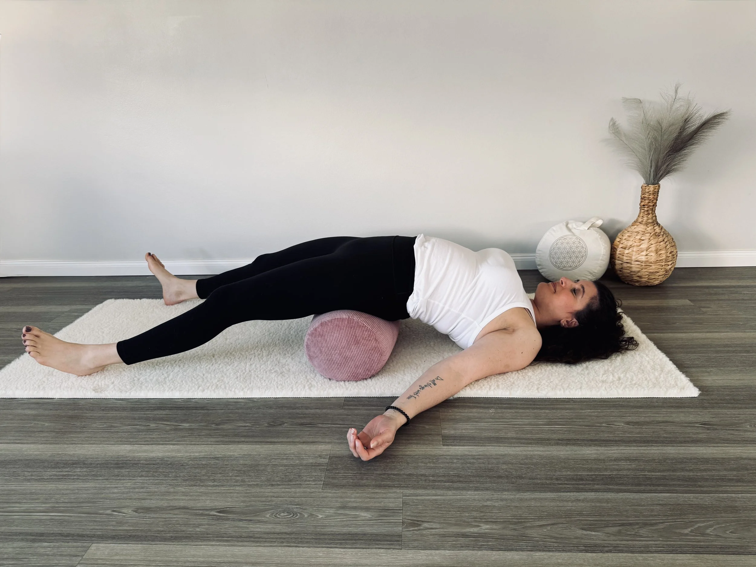 Frau liegt auf der Seite auf einem Yoga-Bolster, übt eine Dehnung mit den Beinen nach oben, im Hintergrund eine weiße Wand mit einem Korb und einem Dekokissen.