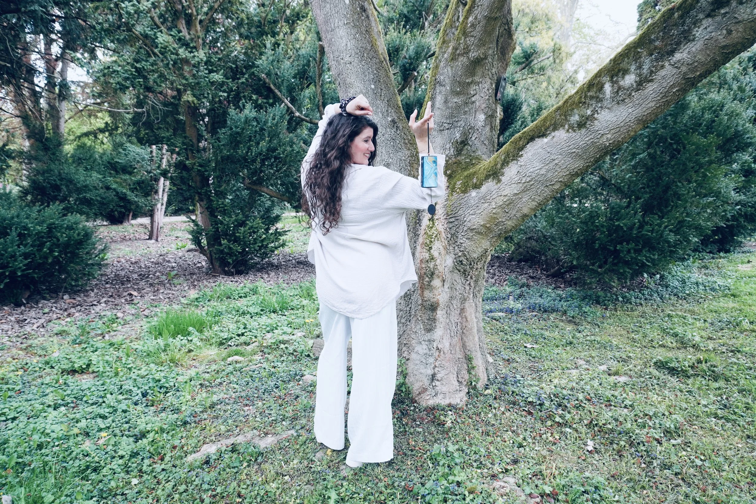 Frau mit langen, lockigen Haaren, weißem Oberteil und weißen Hosen, macht ein Selfie vor einem Baum im grünen Park, während sie eine Smartphone-Hülle trägt.