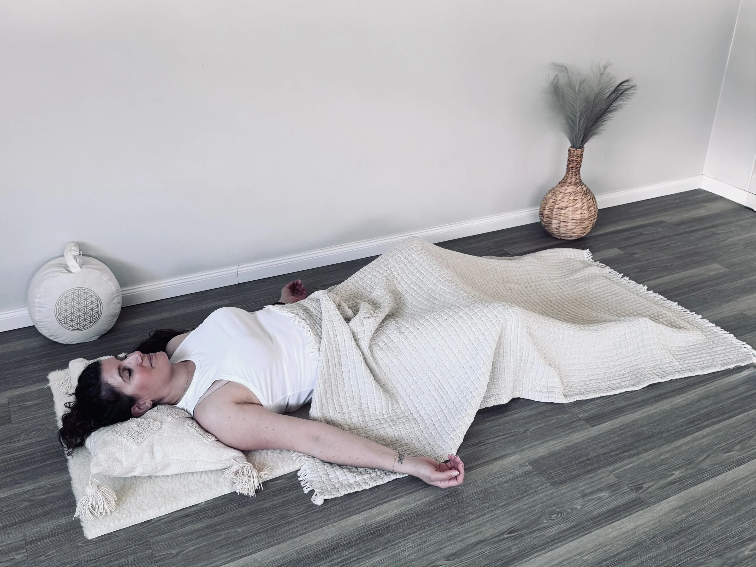 Eine Frau liegt entspannt auf dem Boden mit einem weißen Kissen und einer Decke, die sie bedeckt, neben einer großen Vase mit Federn, im Hintergrund ein weißer Wandabschnitt.