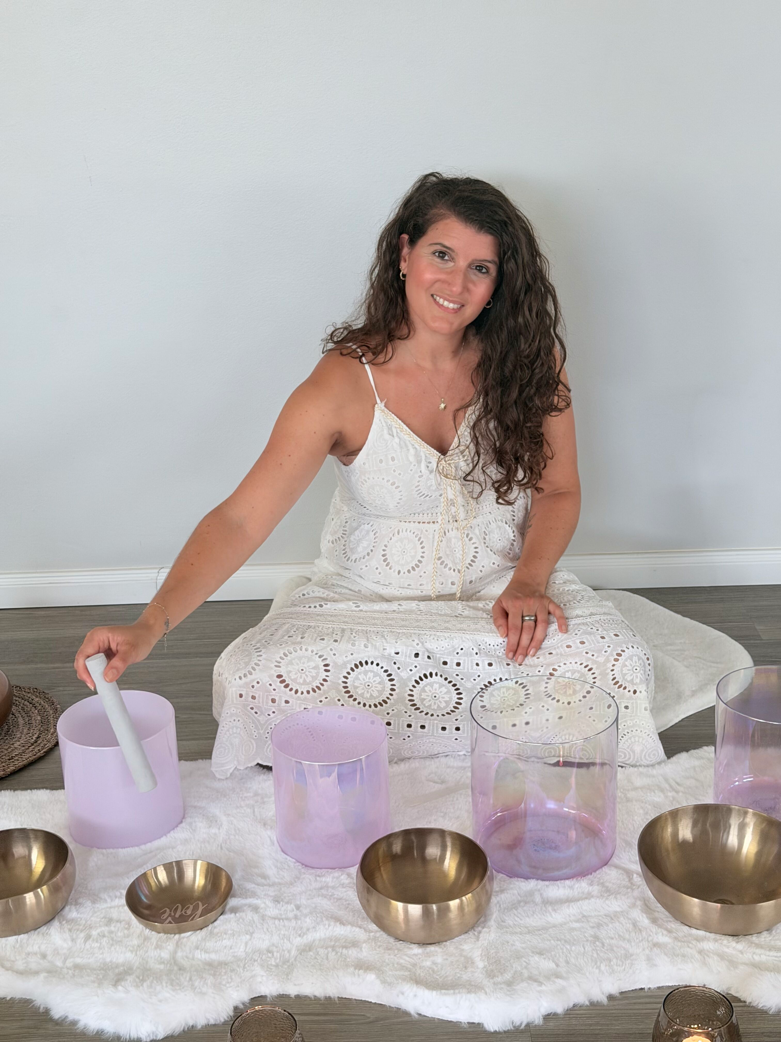 Eine Frau sitzt auf einem weißen Teppich vor einem weißen Hintergrund und spielt auf Kristallklangschalen, umgeben von verschiedenen farbigen Gläsern und Kerzen, was eine Meditations- oder Klangheilungssitzung vermittelt.