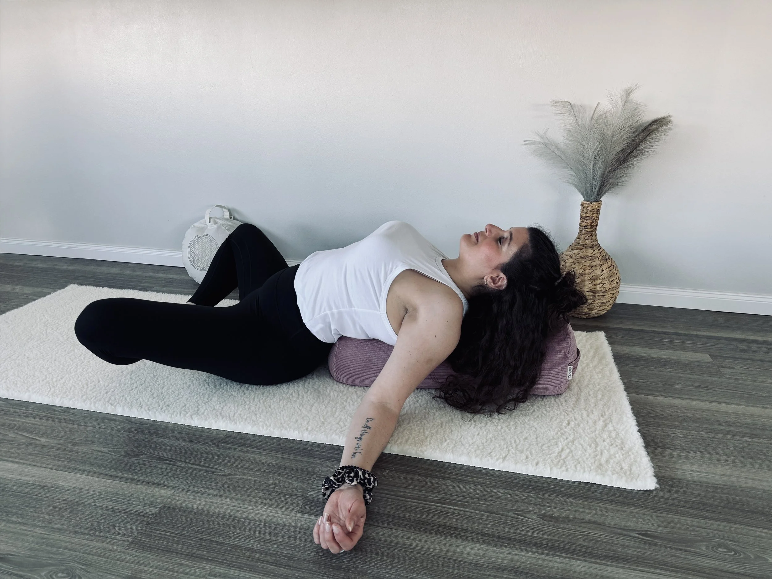 Eine Frau liegt auf dem Rücken auf einem Yogaboden, in einer Schulterbrücke-Position, mit einem Kissen unter ihrem Rücken, während sie eine Übung macht. Der Raum hat weiße Wände und dunkles Holzparkett, mit einem großen, geflochtenen Korb mit getrock