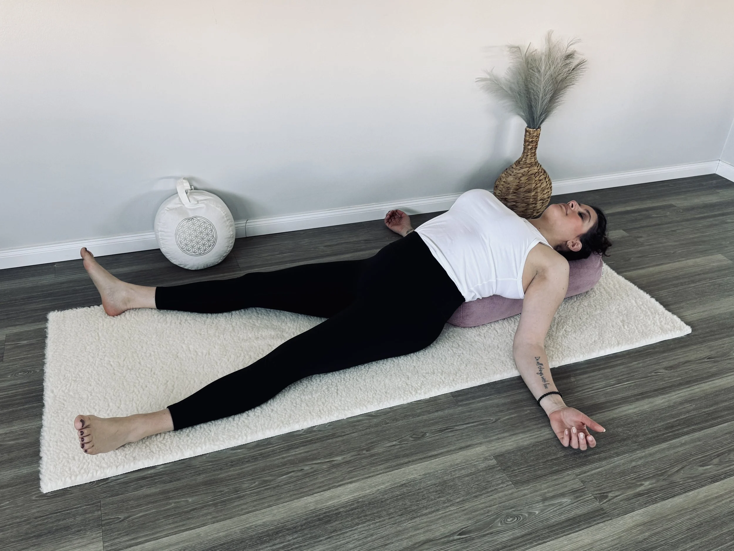 Eine Frau liegt entspannt auf einem Yogamatte, mit den Armen und Beinen ausgestreckt, in einem Raum mit weißen Wänden, neben einer großen Vase mit Federn und einem Gegenstand mit einem geometrischen Muster, im Hintergrund.