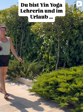 Eine Frau steht auf einem gepflasterten Bereich im Garten, umgeben von grünen Büschen und Bäumen. Sie trägt eine weiße top und schwarze Shorts. Es gibt einen Text-Overlay, der sagt: 'Du bist Yin Yoga Lehrerin und im Urlaub ...'.