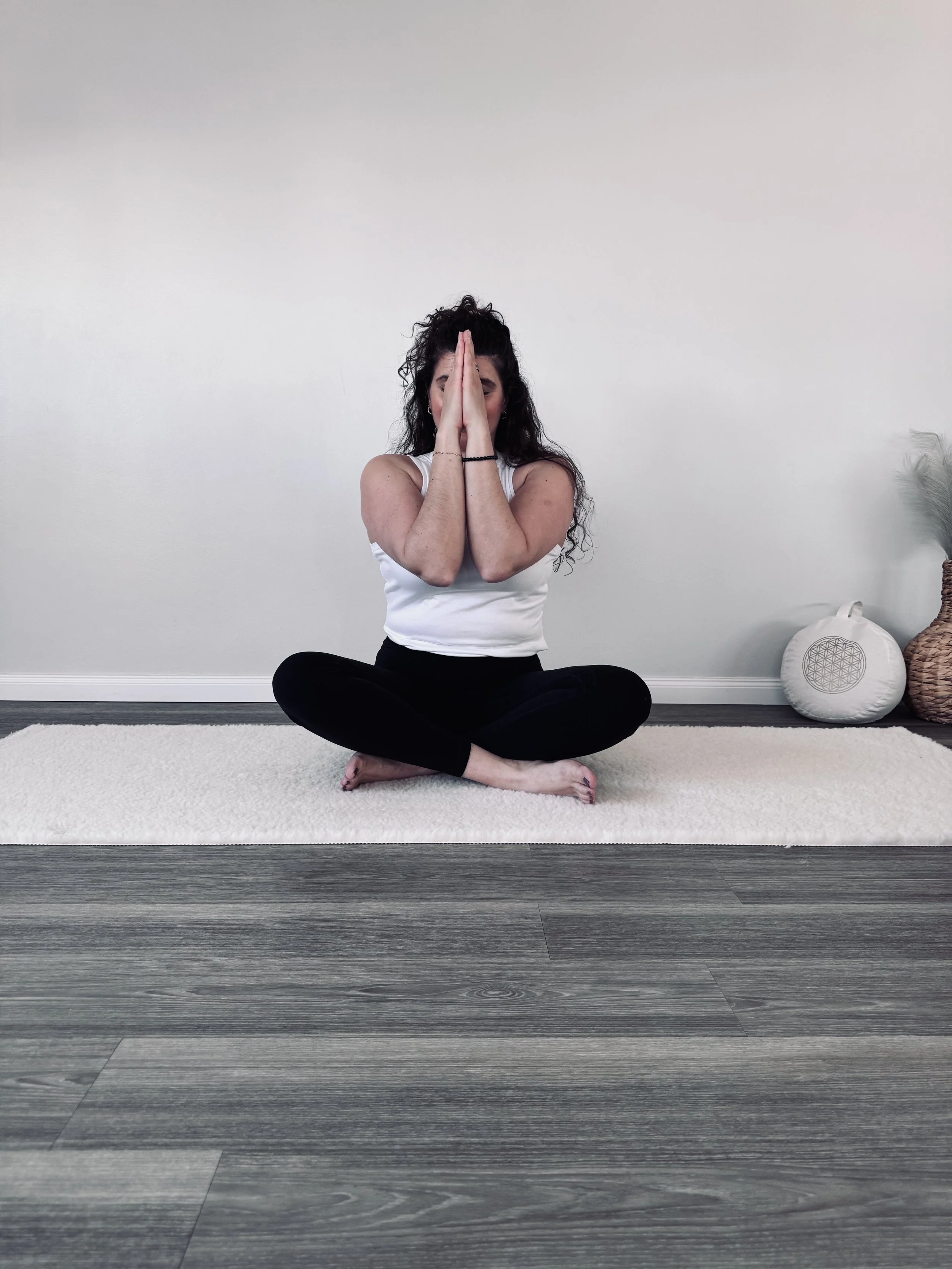 Junge Frau sitzt im Schneidersitz auf einem weißen Teppich vor einer weißen Wand und macht eine Yoga-Geste mit den Händen vor dem Gesicht. Sie trägt ein weißes Tanktop und schwarze Hosen.
