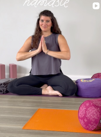 Eine Frau sitzt in einem Yoga-Haltung, die Hände in Gebetshaltung vor der Brust. Sie befindet sich in einem Yoga-Studio mit Yogamatten und Kissen im Hintergrund.