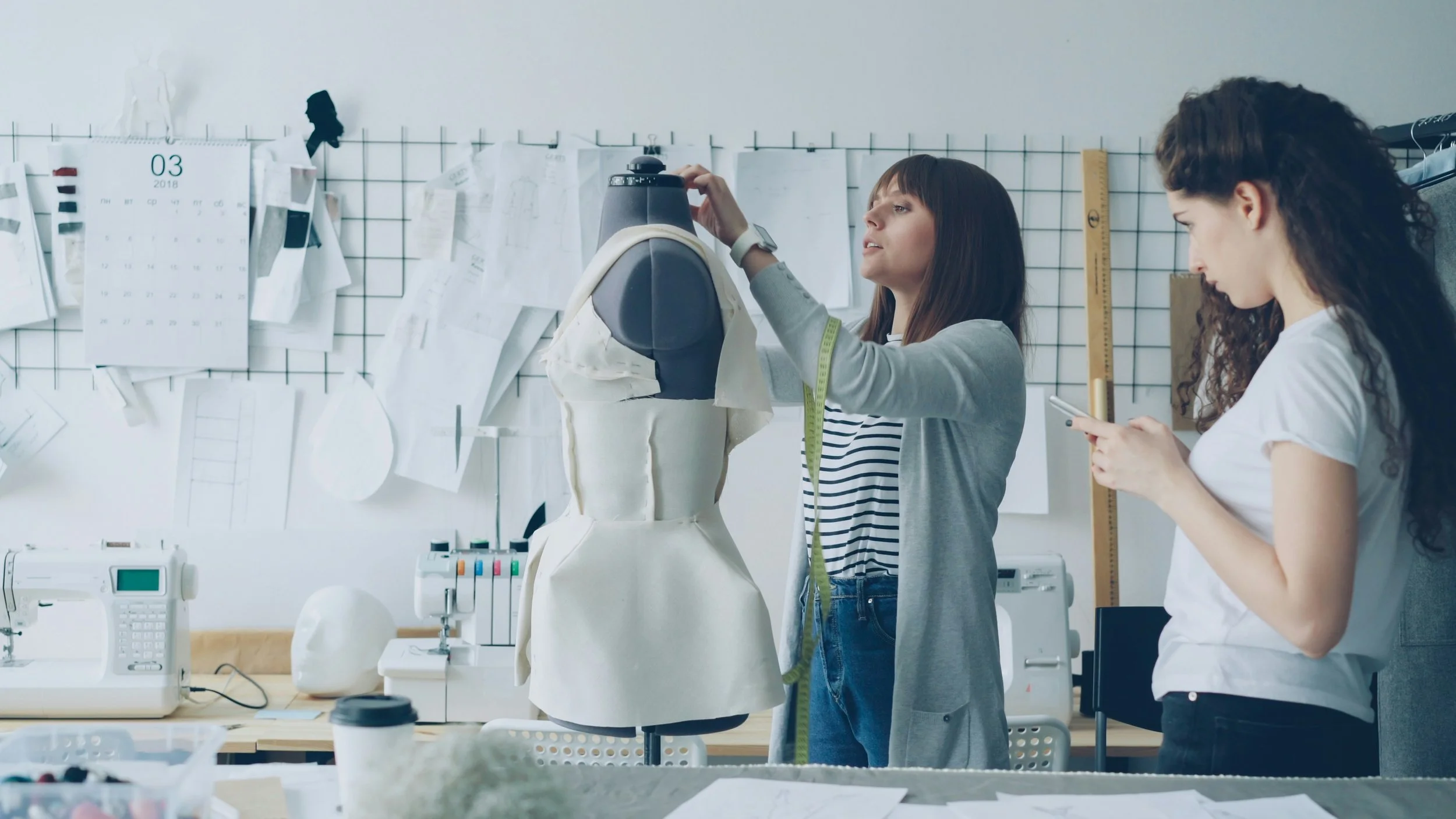 Två kvinnor arbetar i en designstudio, en justerar en kläddocka och den andra använder sin mobil.