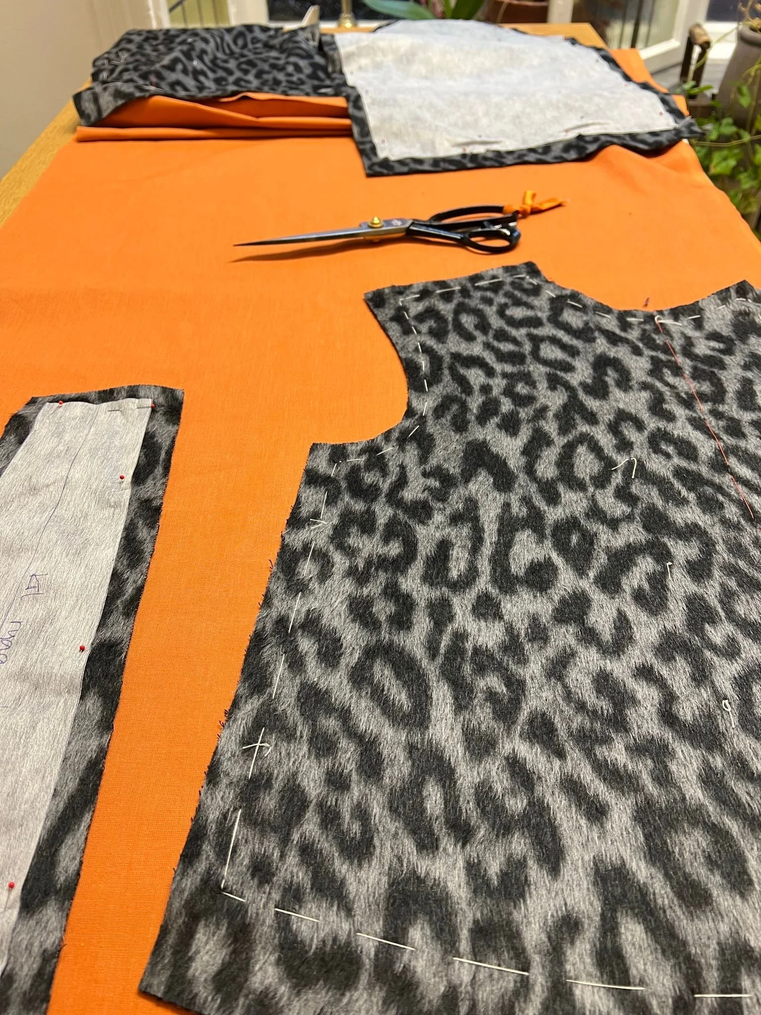 Textiltyger med leopardmönster och orange tyg styckas på ett bord, med ett par saxar och nålar.