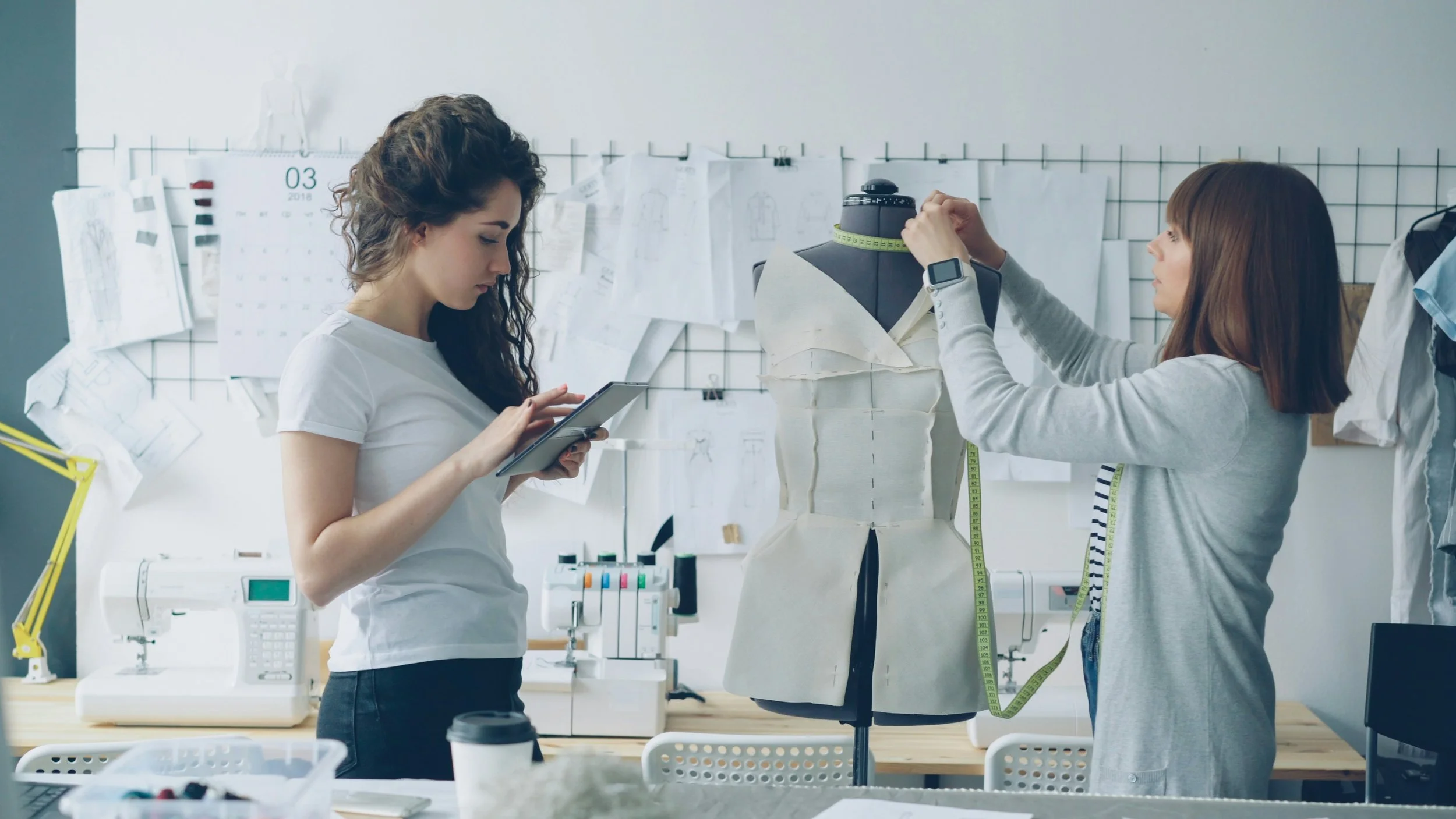 Två kvinnor arbetar i en designstudio. Den ena granskar en mönsterdocka med måttband, den andra tittar på sin telefon. Bakom finns ritningar och mönsterklipp på väggarna.