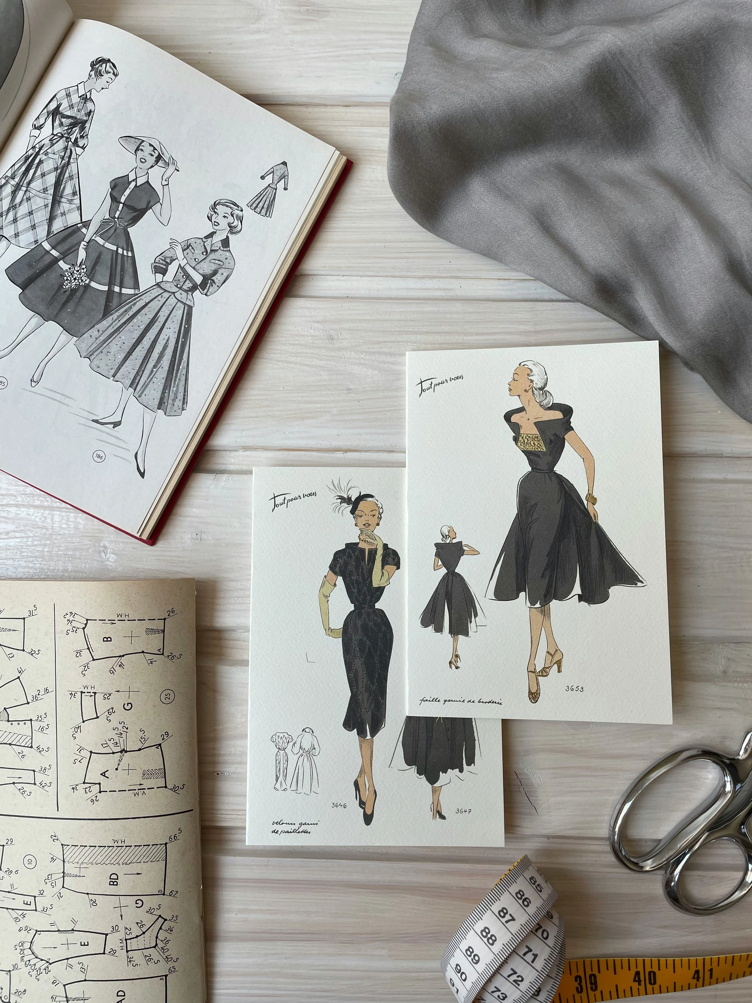 Illustrations av kvinnor i eleganta kläder, mönsterritningar för kläder, mätskivor och textilplaner på ett skrivbord.