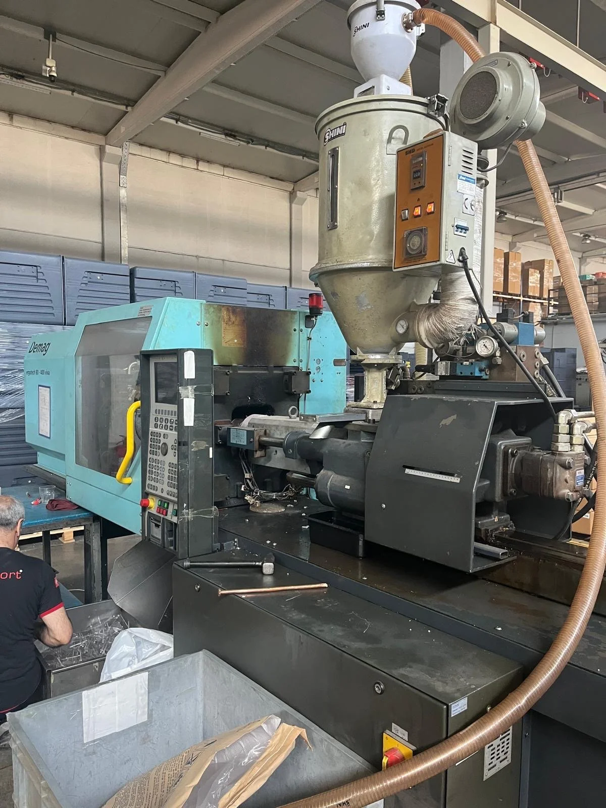 Industrial manufacturing machinery with a large vertical metal hopper and a control panel, situated in a factory setting.