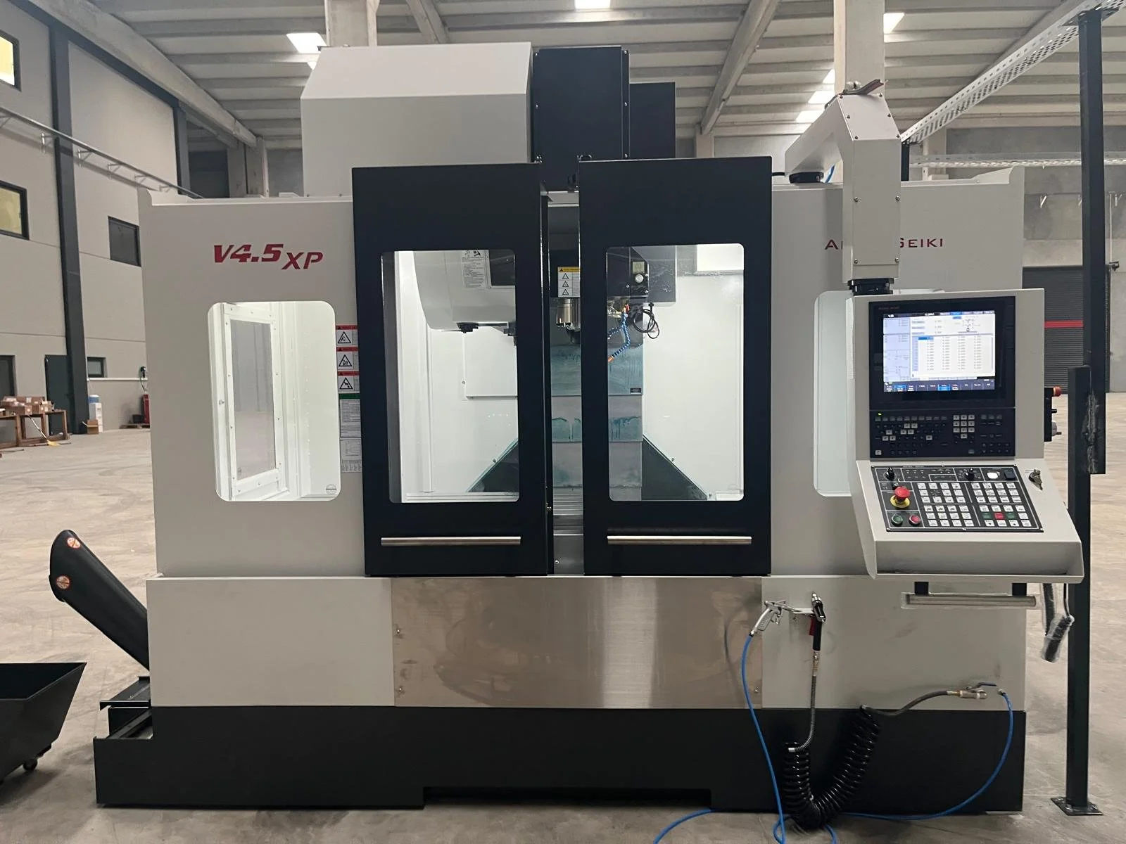 A computer numerical control (CNC) milling machine with a control panel on the right side featuring a monitor, keypad, and emergency stop button, located in an industrial warehouse setting.