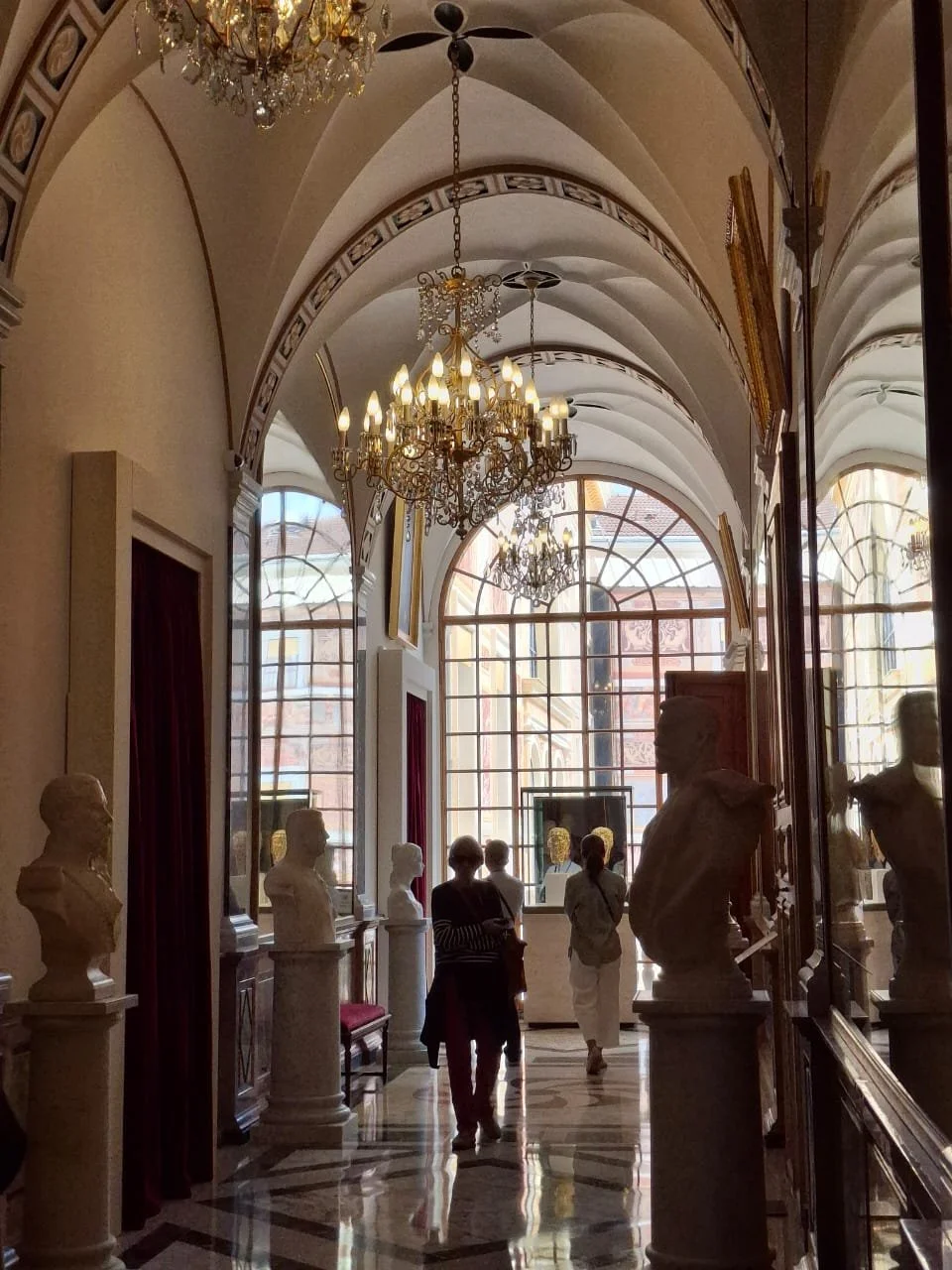Interior of a museum or gallery with bust sculptures on pedestals along the sides, ornate chandeliers hanging from the ceiling, large arched windows letting in natural light, and visitors walking through.