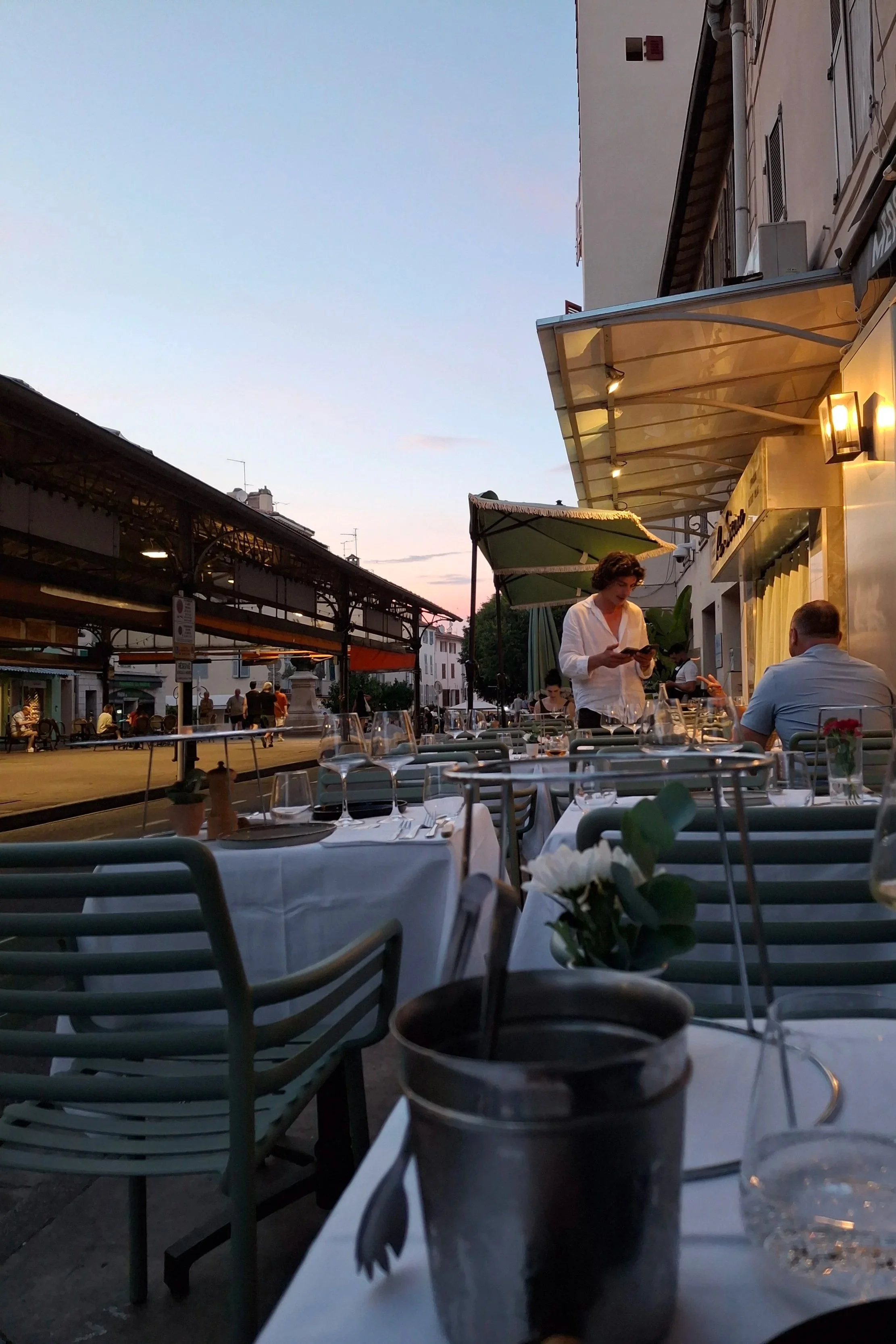 Outdoor restaurant scene at dusk with tables set with glasses and flowers, people dining, a waitress taking an order, and a building with a balcony and hanging lights in the background.