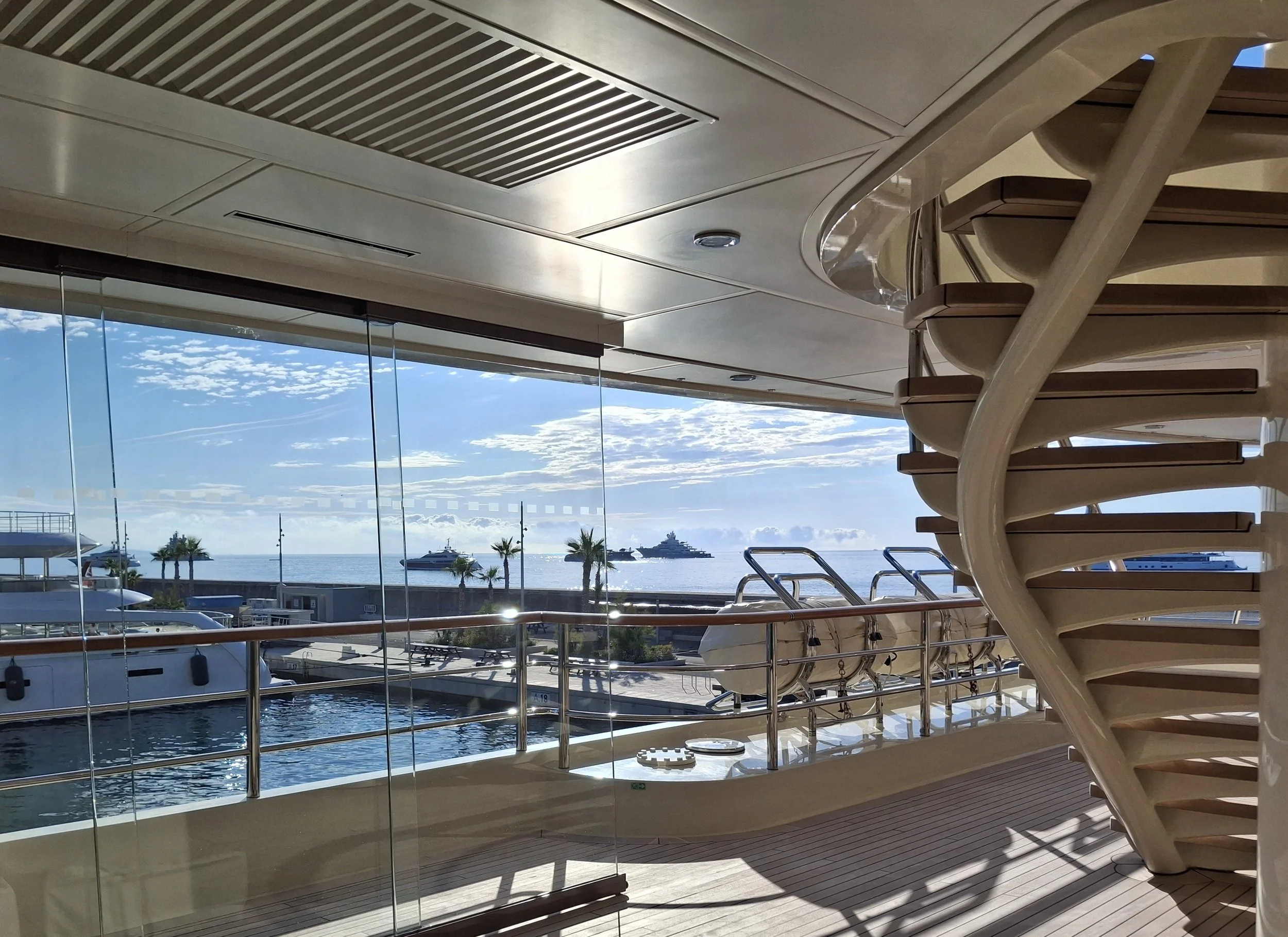 View from a balcony of a marina with boats and yachts, overlooking the ocean with a cruise ship in the distance, under a partly cloudy sky.