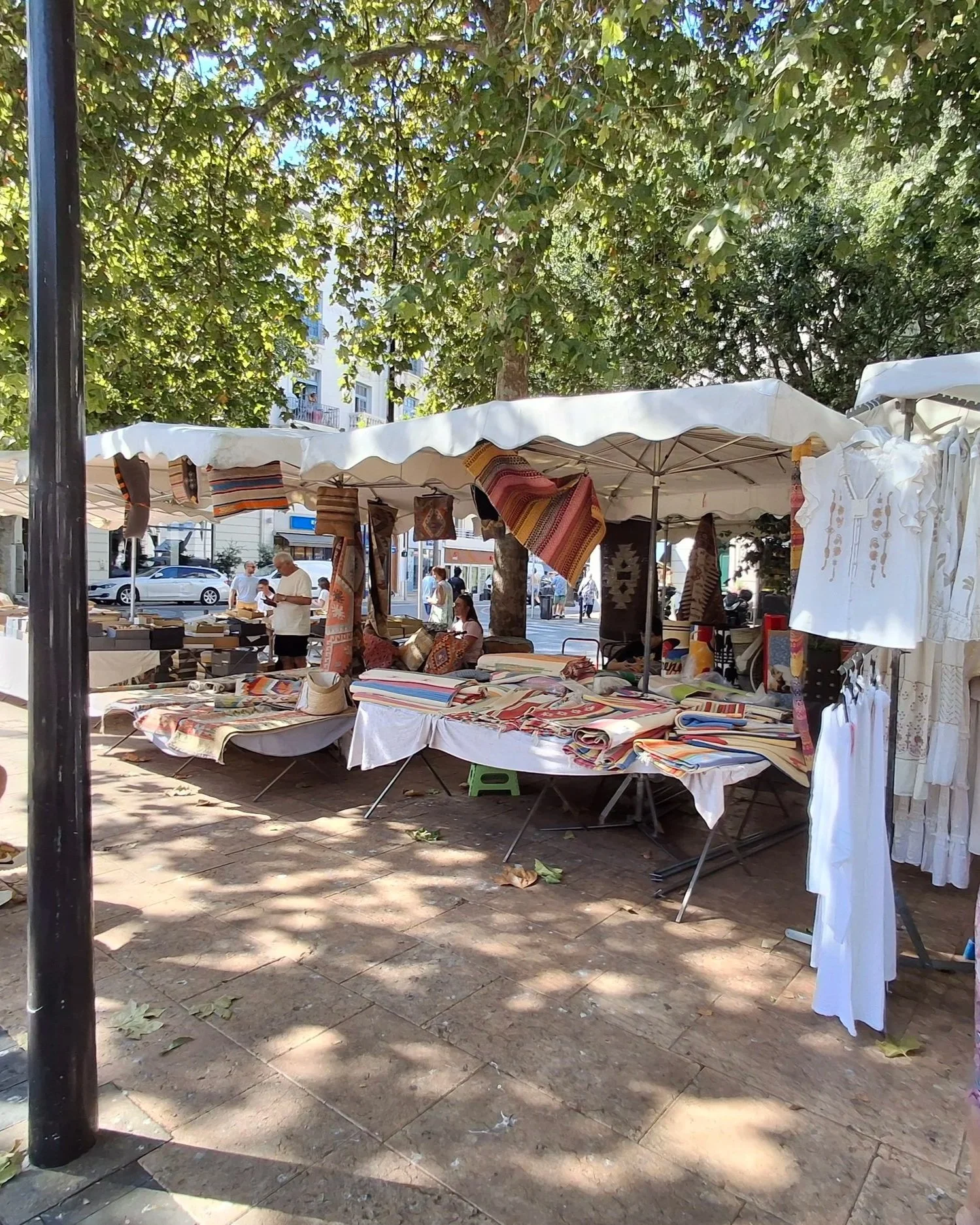 An outdoor market stall under a large tree, displaying various textiles and clothing, with a white canopy and trees casting shadows on the ground. Several people are visible, shopping and browsing.