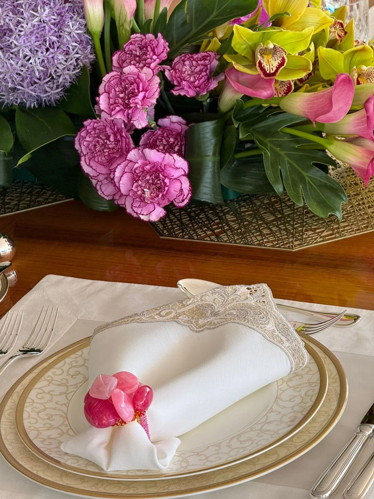 A formal table setting features a white napkin with embroidered lace trim, decorated with pink heart-shaped candies and a pink plastic flower. The table is set with elegant gold-rimmed plates, silver utensils, and a white tablecloth. Behind the table, a floral arrangement of pink carnations, yellow calla lilies, pink and cream orchids, and purple hyacinths is visible, placed on a wooden surface.