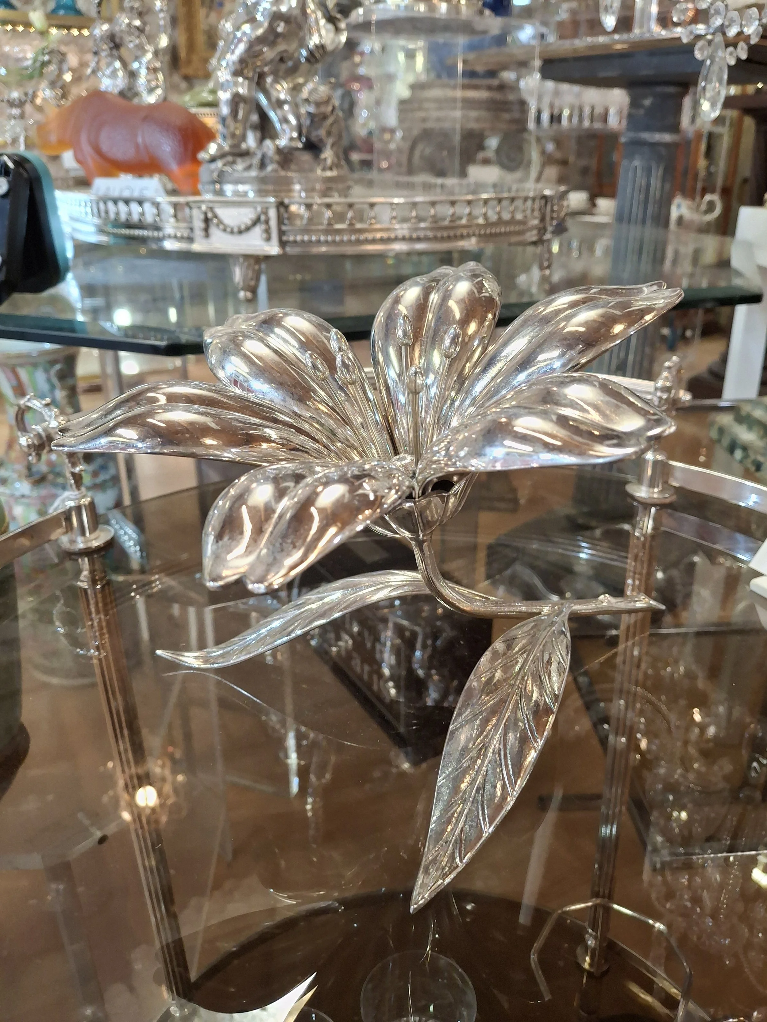 A decorative glass flower with elongated petals, placed on a reflective surface. In the background, there are various ornate items including a silver tray, a ceramic figure, and other decorative objects.