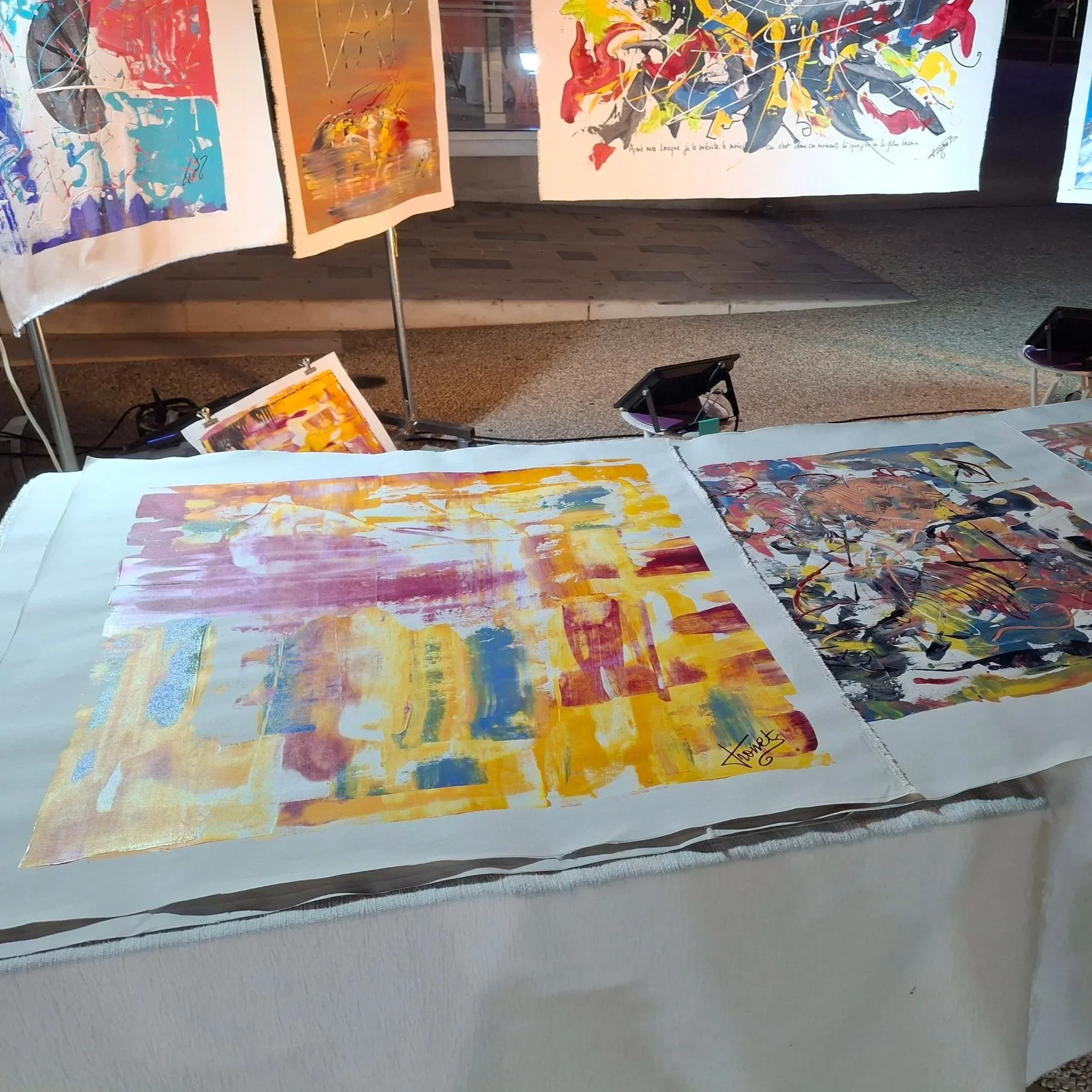 Multiple colorful modern abstract paintings displayed at an art exhibition, with splashes, strokes, and mixed media on paper or canvas, signed by the artist.