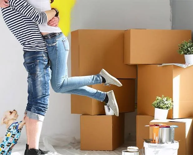 A couple standing in front of a wall with multiple stacked cardboard boxes, one person is lifting another person by the legs while they hold hands, a small dog with a backpack looks up at them, and there are plants and painting supplies on the floor.