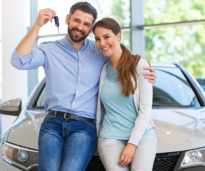 A happy couple standing beside a silver car in a dealership, holding car keys, with the man smiling and the woman leaning into him.