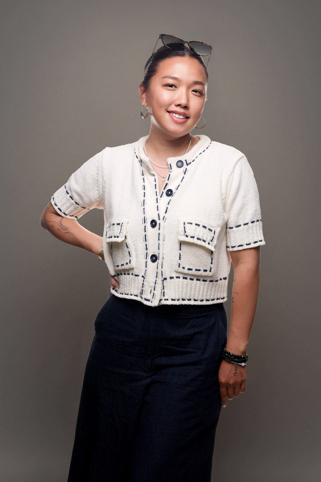 A young woman with dark hair, wearing sunglasses on her head, earrings, a cream-colored short-sleeved jacket with pockets, and dark pants, smiling with one hand on her hip against a plain gray background.