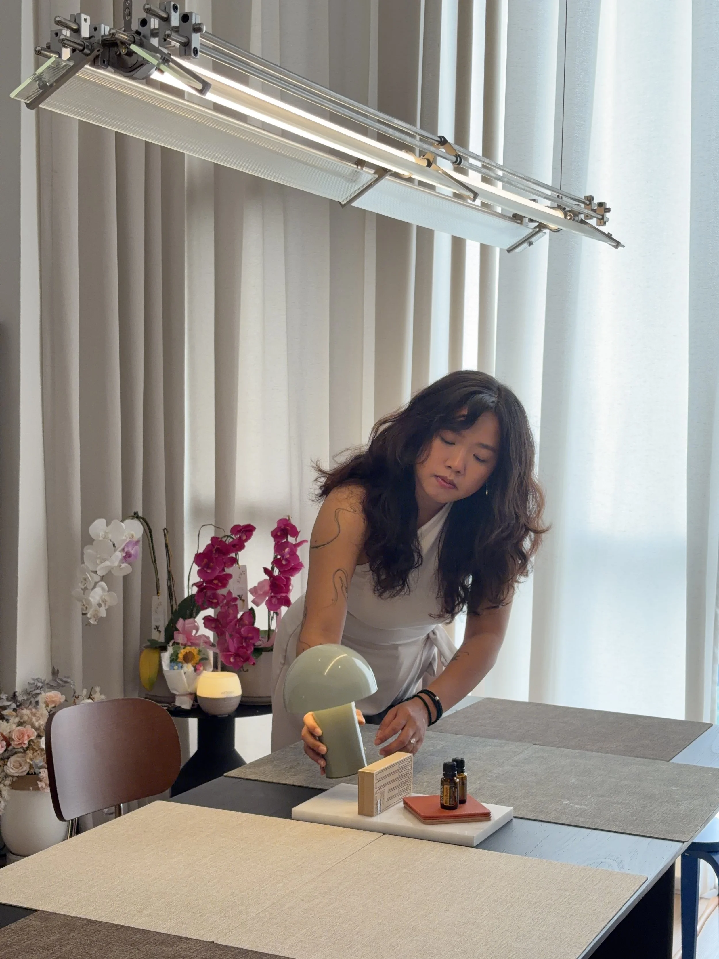 Woman arranging essential oils and a diffuser on a dining table, with flowers and paper items nearby, in a bright room with curtains.