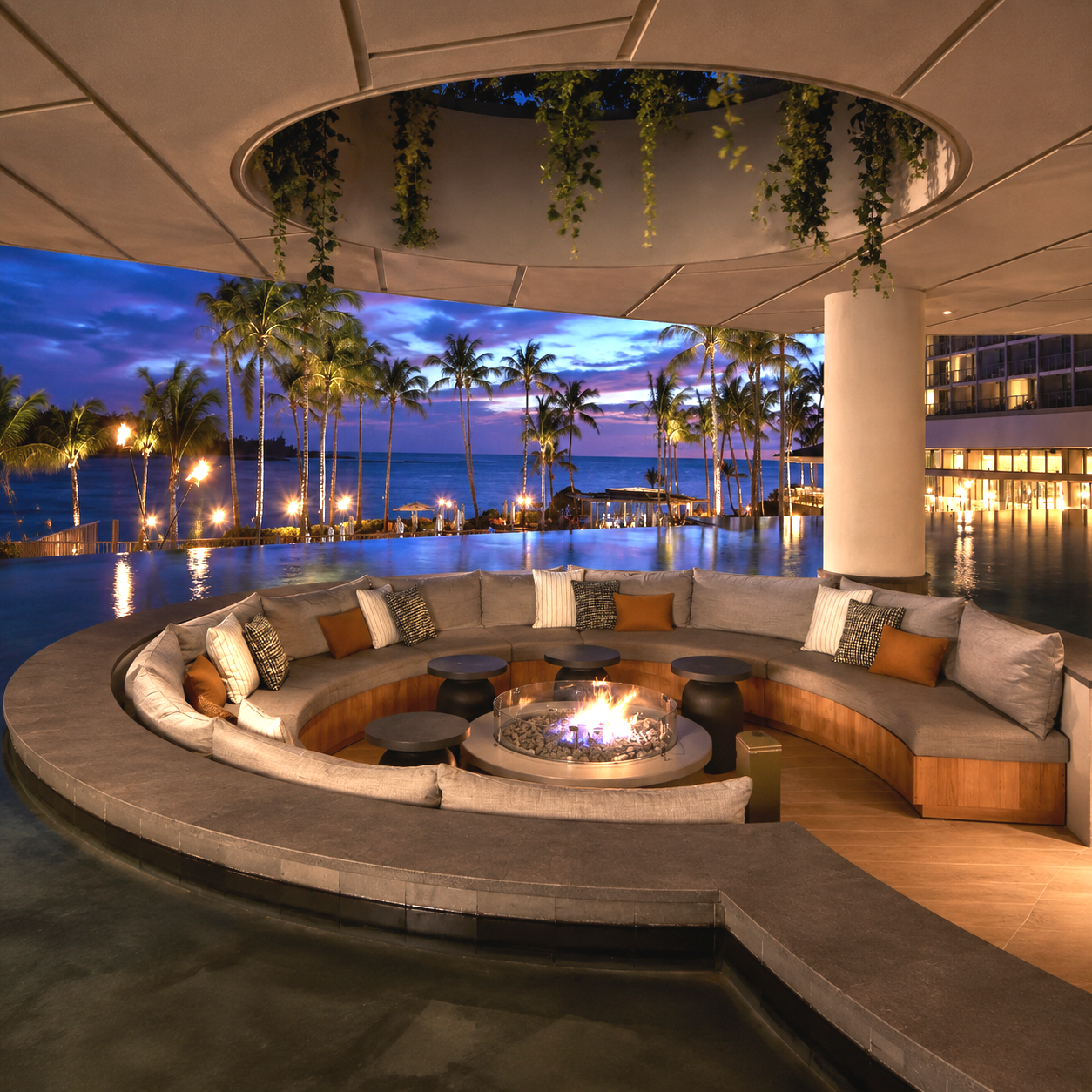 Luxury outdoor lounge area with curved sofa and fire pit, overlooking a tropical beach and palm trees at sunset, with a pool and a large building with balconies in the background.