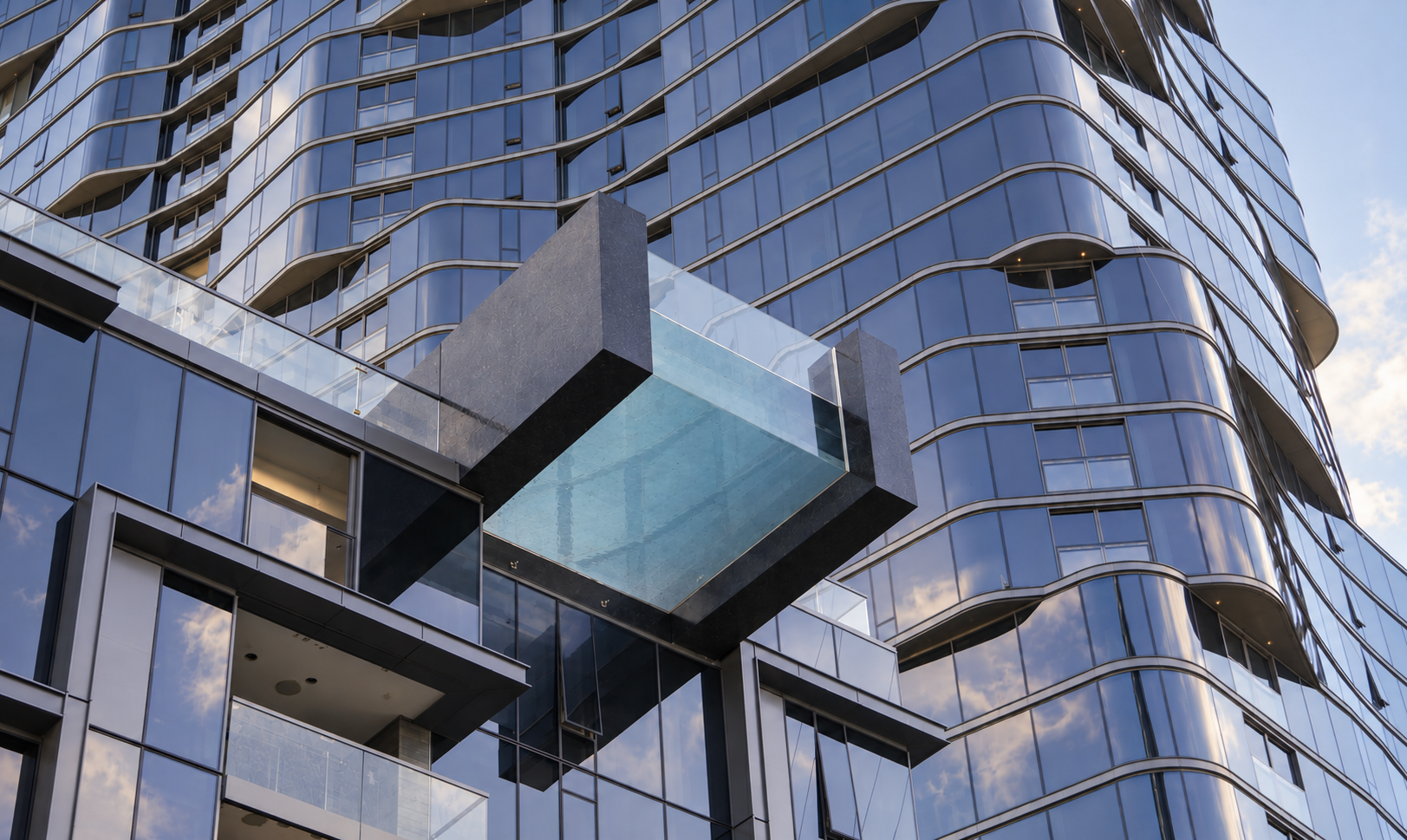 Close-up of a modern, glass skyscraper with a protruding glass balcony and curved windows reflecting the sky and clouds.