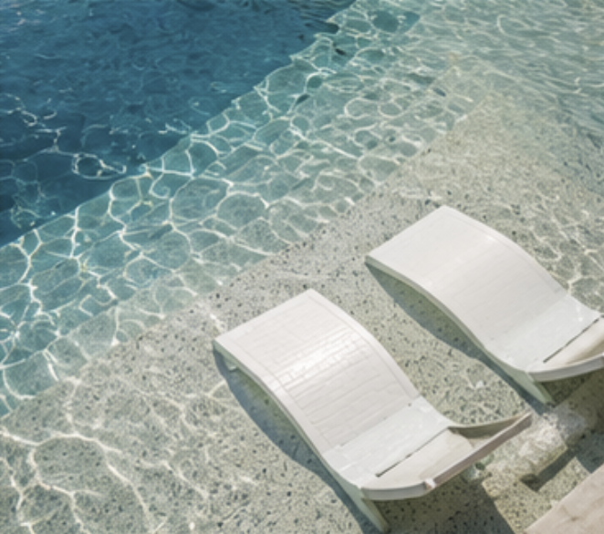 Two white lounge chairs on a poolside with clear blue water.