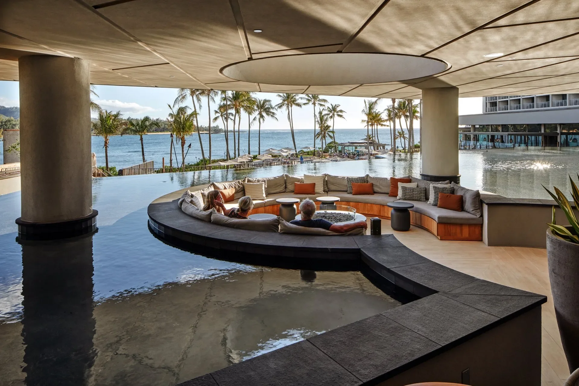 An indoor pool area overlooking a scenic ocean view with palm trees. There is a circular seating area with cushions and a fire pit in the center, and two people are relaxing on the cushions.
