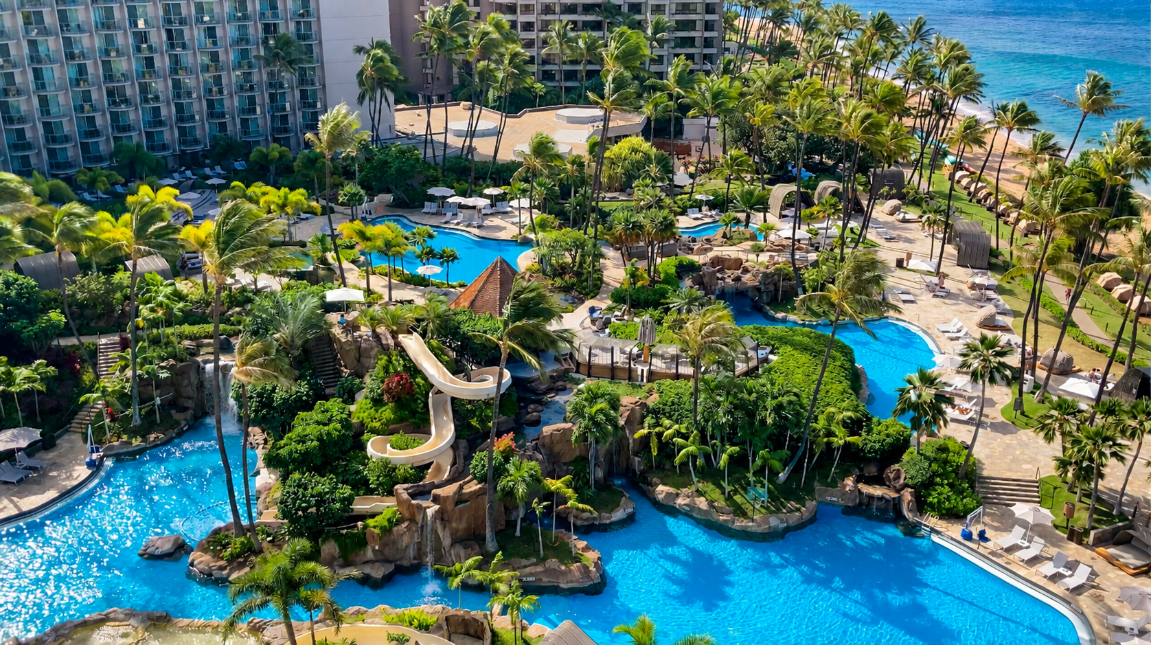Aerial view of a luxury resort with multiple swimming pools surrounded by palm trees, lounge chairs, and lush greenery near the ocean.