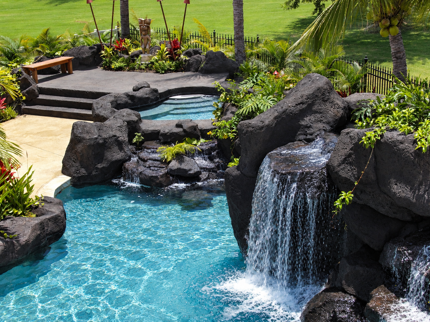 A backyard with a swimming pool featuring a small waterfall and rocks, surrounded by lush tropical plants and greenery, with a grassy lawn and trees in the background.