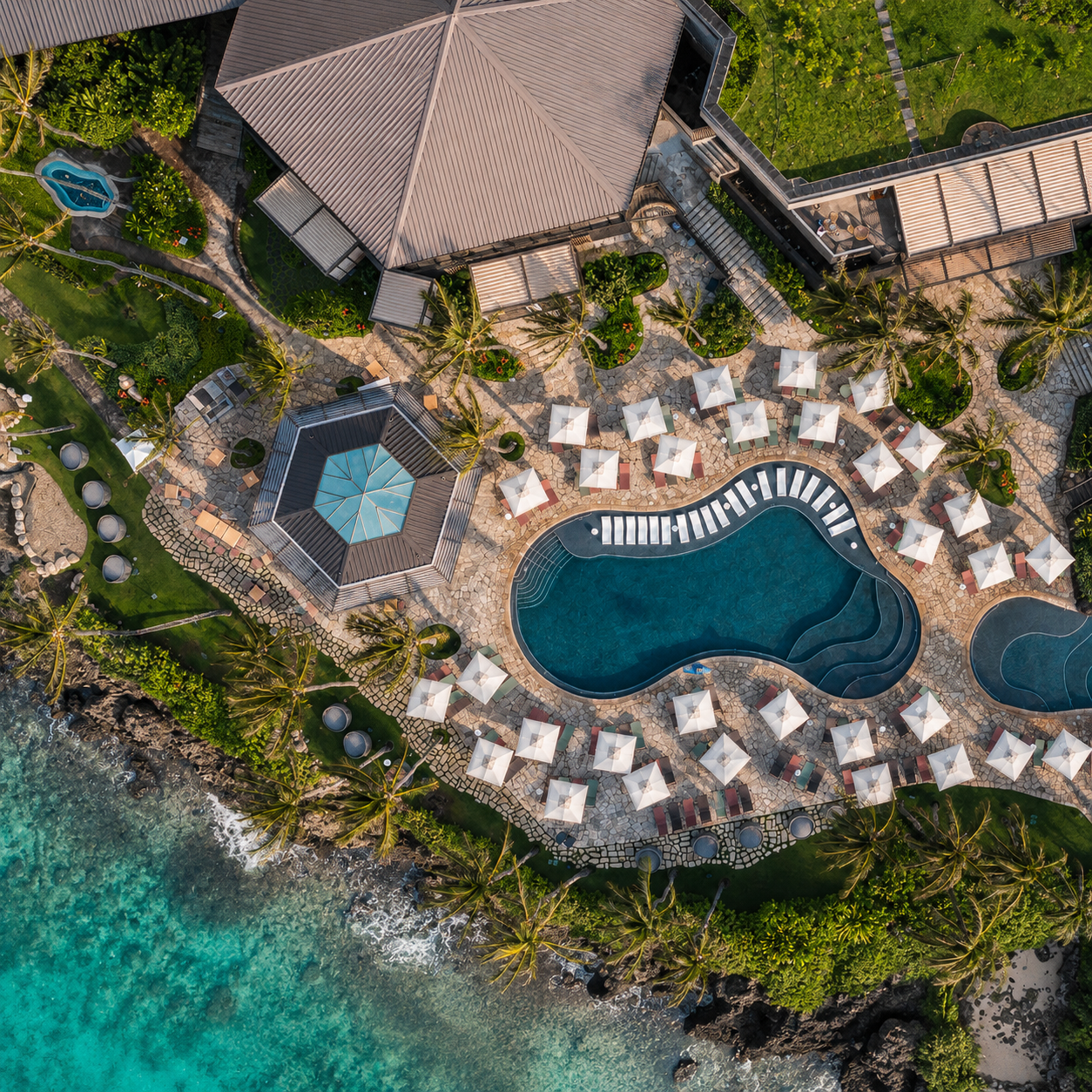 Aerial view of a luxurious tropical resort with a large irregularly shaped swimming pool, surrounded by white umbrellas and lounge chairs, lush green palm trees, a small hut with a lanai, a hot tub, and ocean waves crashing onto the rocky shoreline.