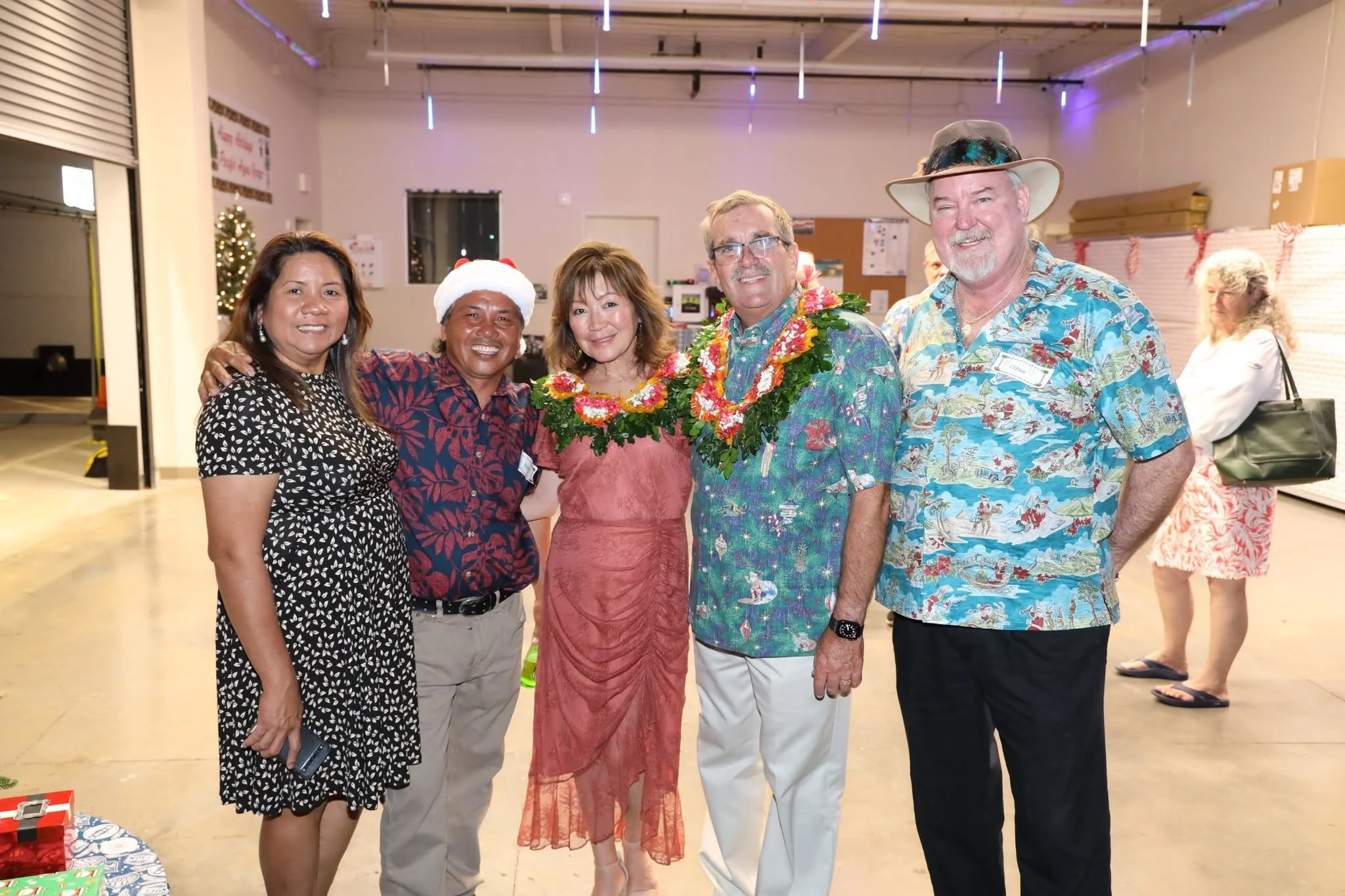 A group of five people smiling and posing together at a festive holiday party. They are dressed in casual and semi-formal attire, with some wearing Hawaiian shirts, a woman in a dress, and a man in a floral shirt with a lei and a hat. The background shows a decorated Christmas tree and holiday decorations.
