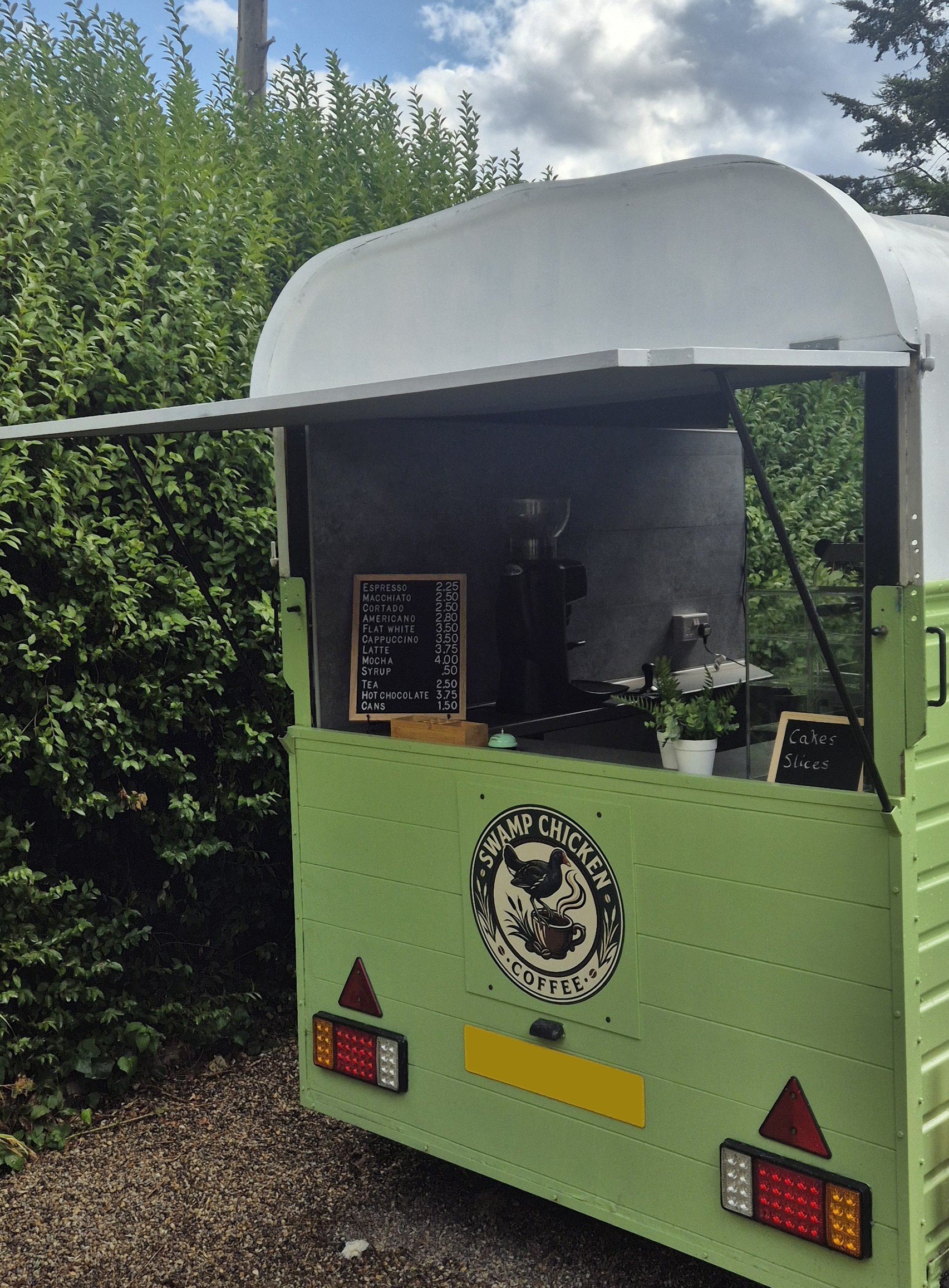 A green food truck selling coffee and drinks, with a menu listing espresso, macchiato, cortado, americano, flat white, cappuccino, latte, mocha, syrup, tea, hot chocolate, and cans, parked outdoors among green bushes and trees.