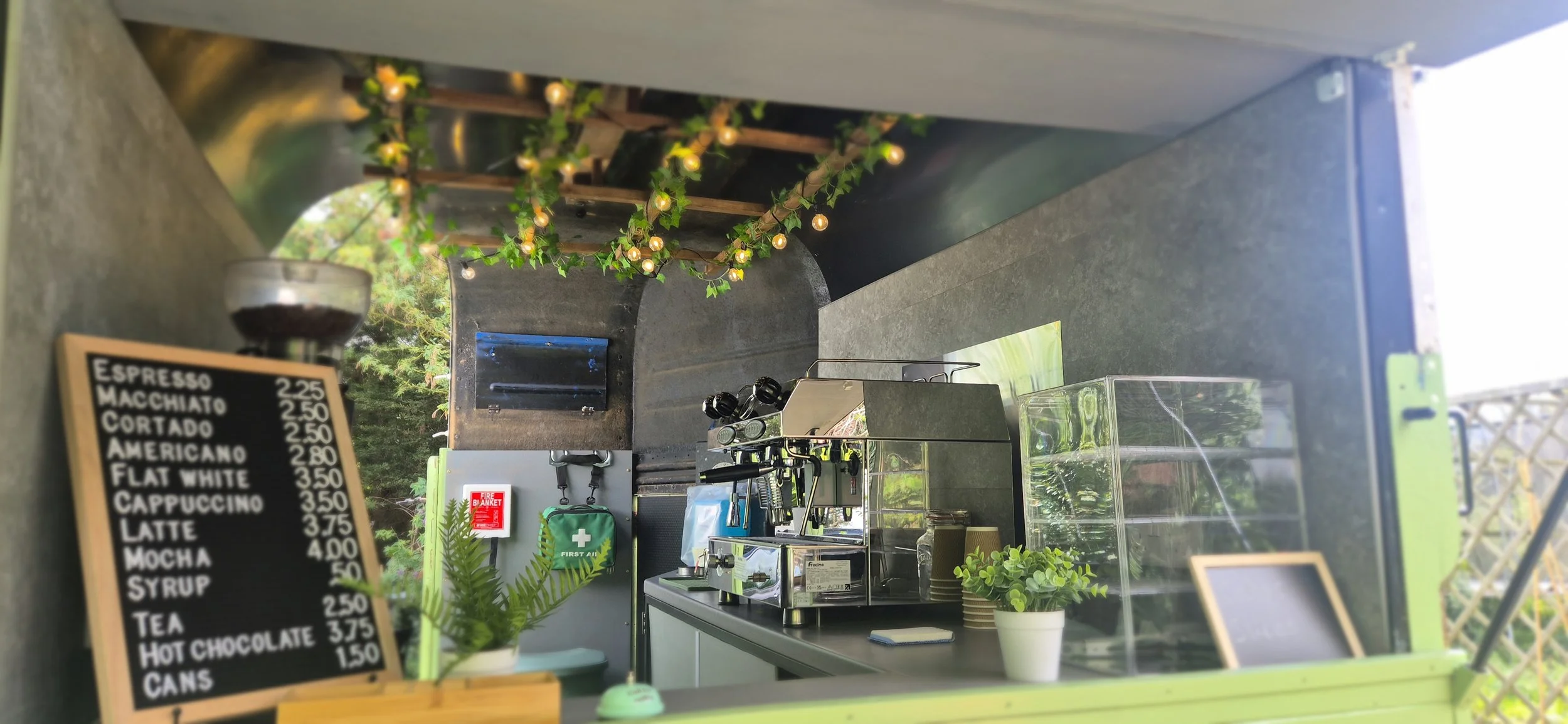 A coffee stand with a blackboard menu featuring various coffee drinks and prices, surrounded by decorative string lights, greenery, and outdoor trees.