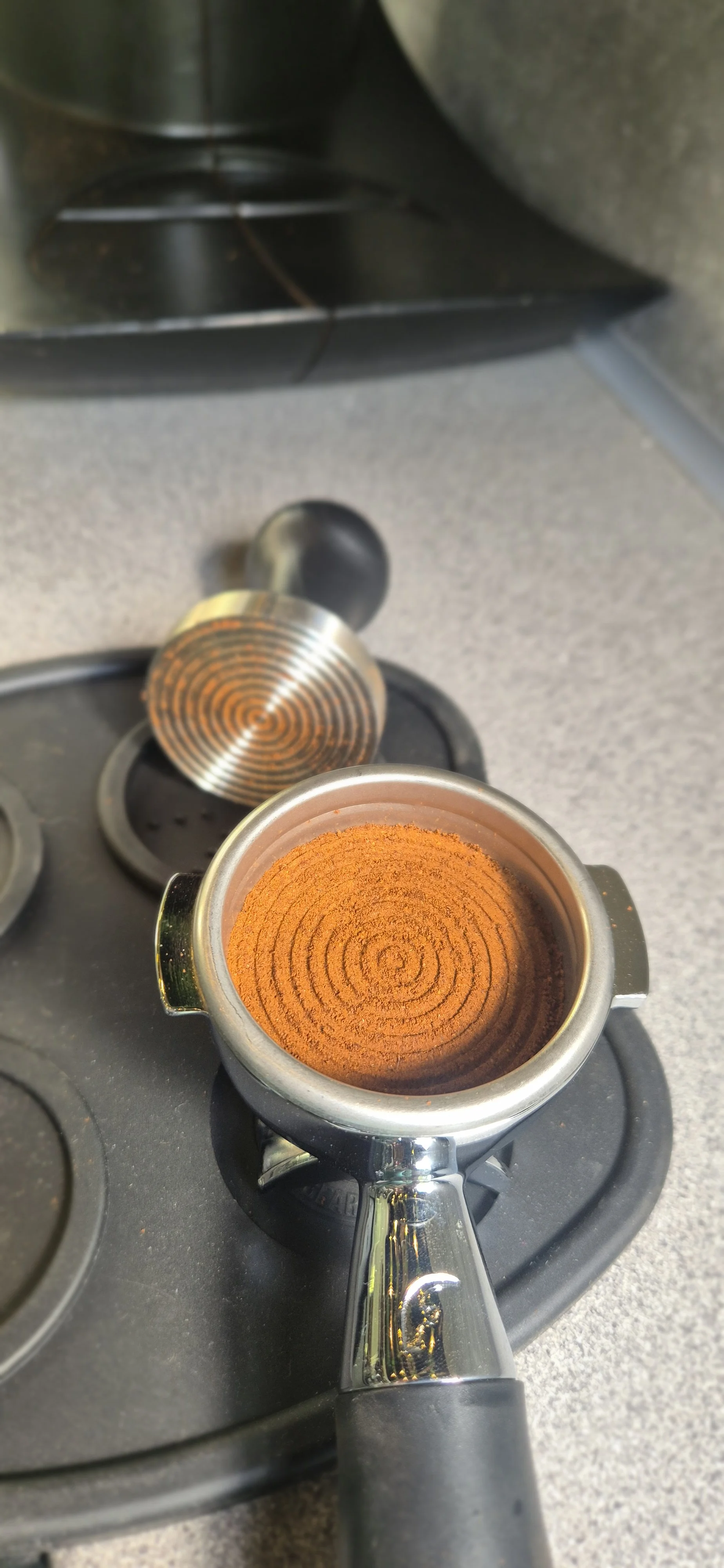 A portafilter filled with finely ground coffee sits on a countertop, with a knock box and a tar color coffee tamper in the background.