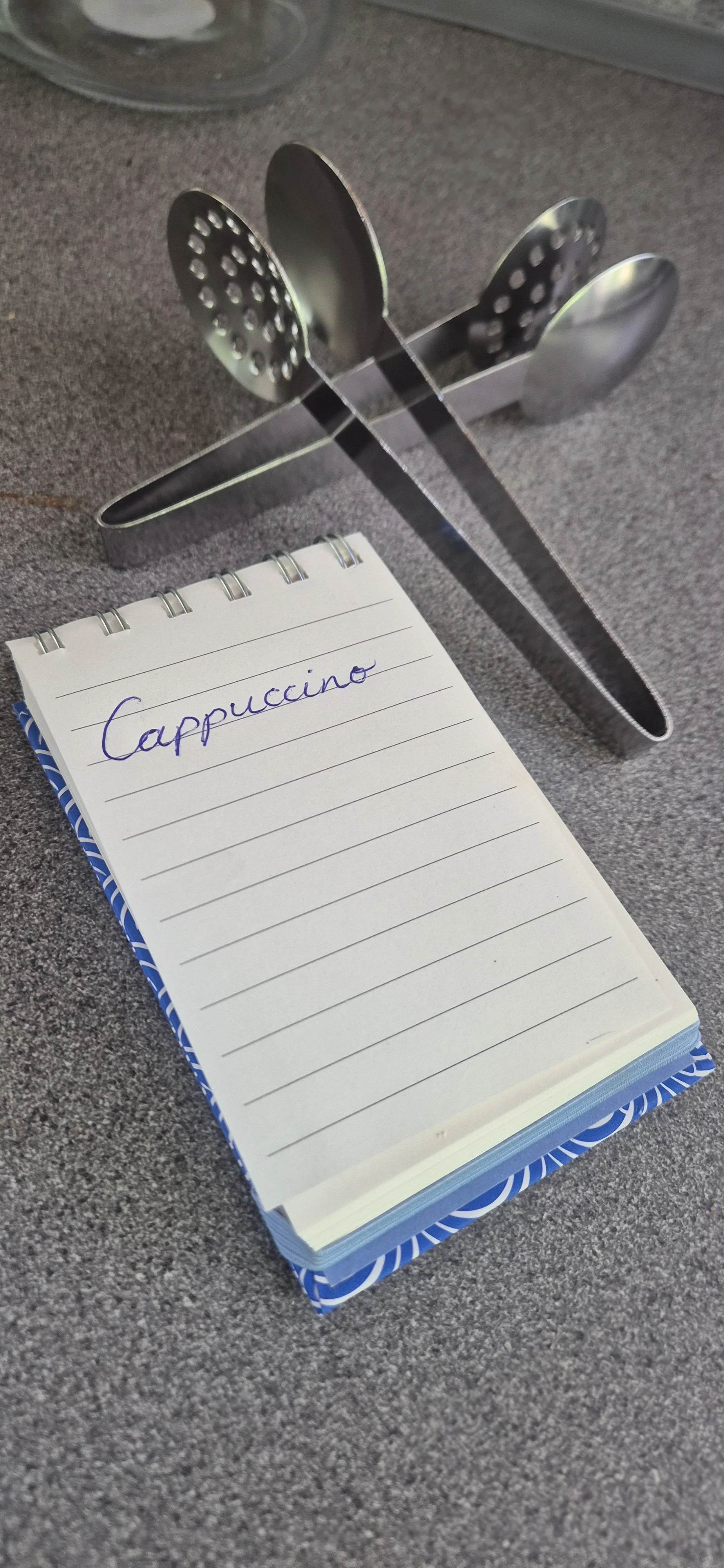 A notebook with the word 'Cappuccino' handwritten on the first page, placed on a gray surface, with metal kitchen utensils with perforated heads behind it.